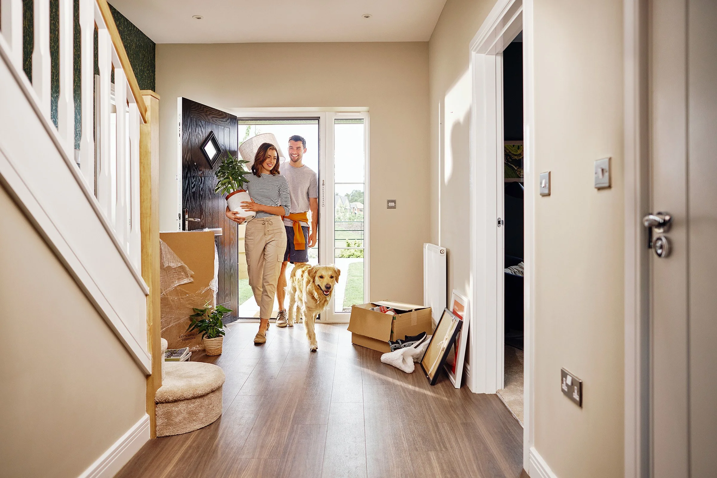 A woman and man entering the house with a golden retriever dog, standing near the doorway. The woman is holding a potted plant. There are boxes and framed pictures on the floor inside the house.