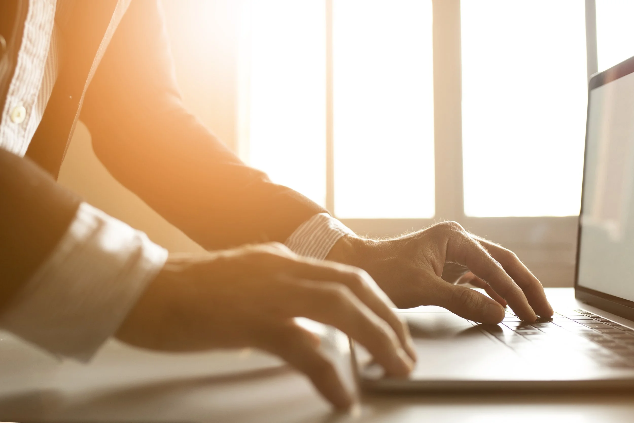 Person typing on a laptop with sunlight coming through a window in the background.