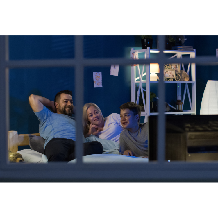 Family of three, two adults and a child, watching television from their bed in a cozy, dimly lit bedroom at night.