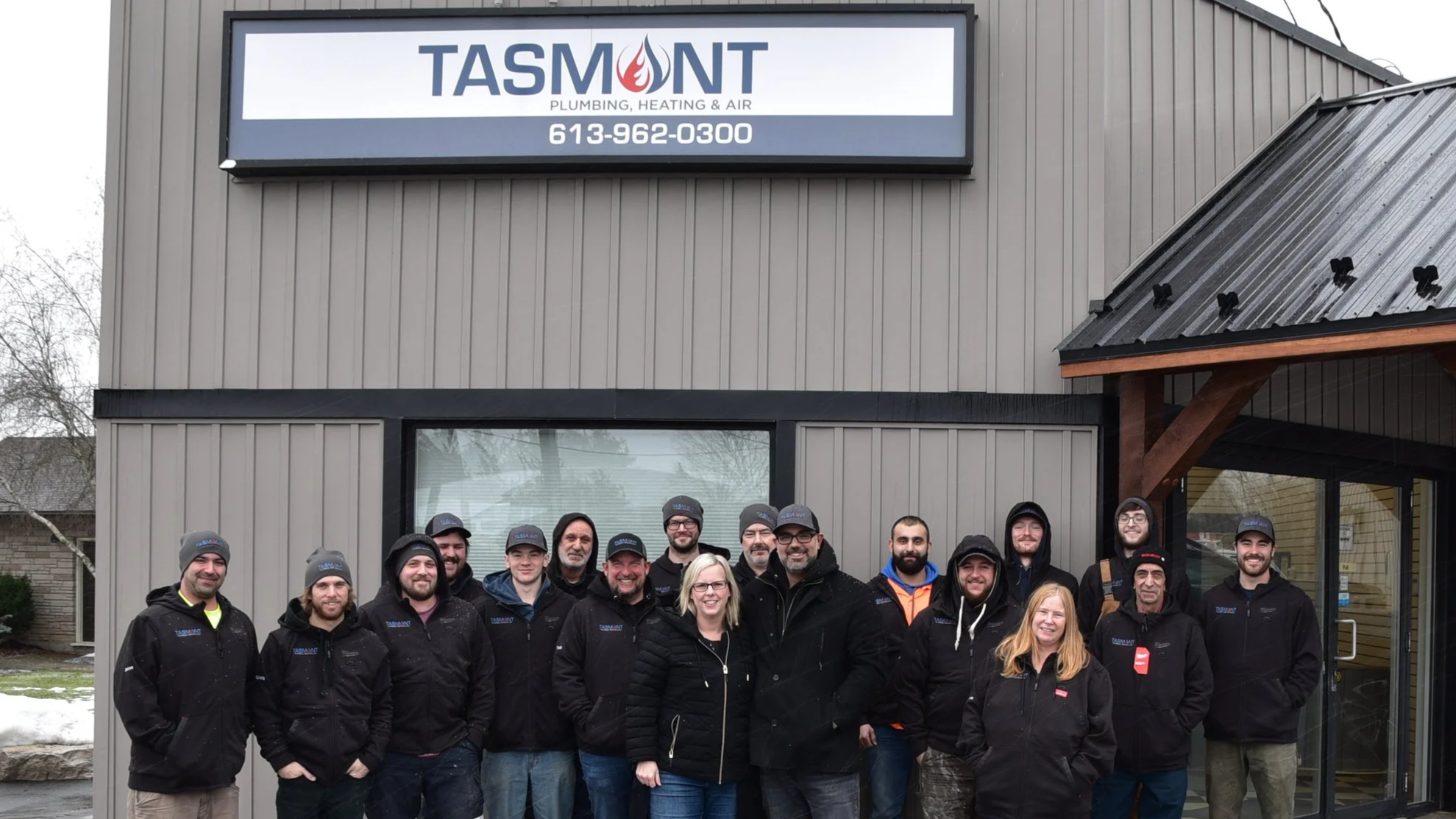 Group of people standing outside a building with a sign that reads TASMONT Plumbing, Heating & Air, with a phone number, wearing dark jackets and hats, some smiling, in front of a gray building with a metal roof.