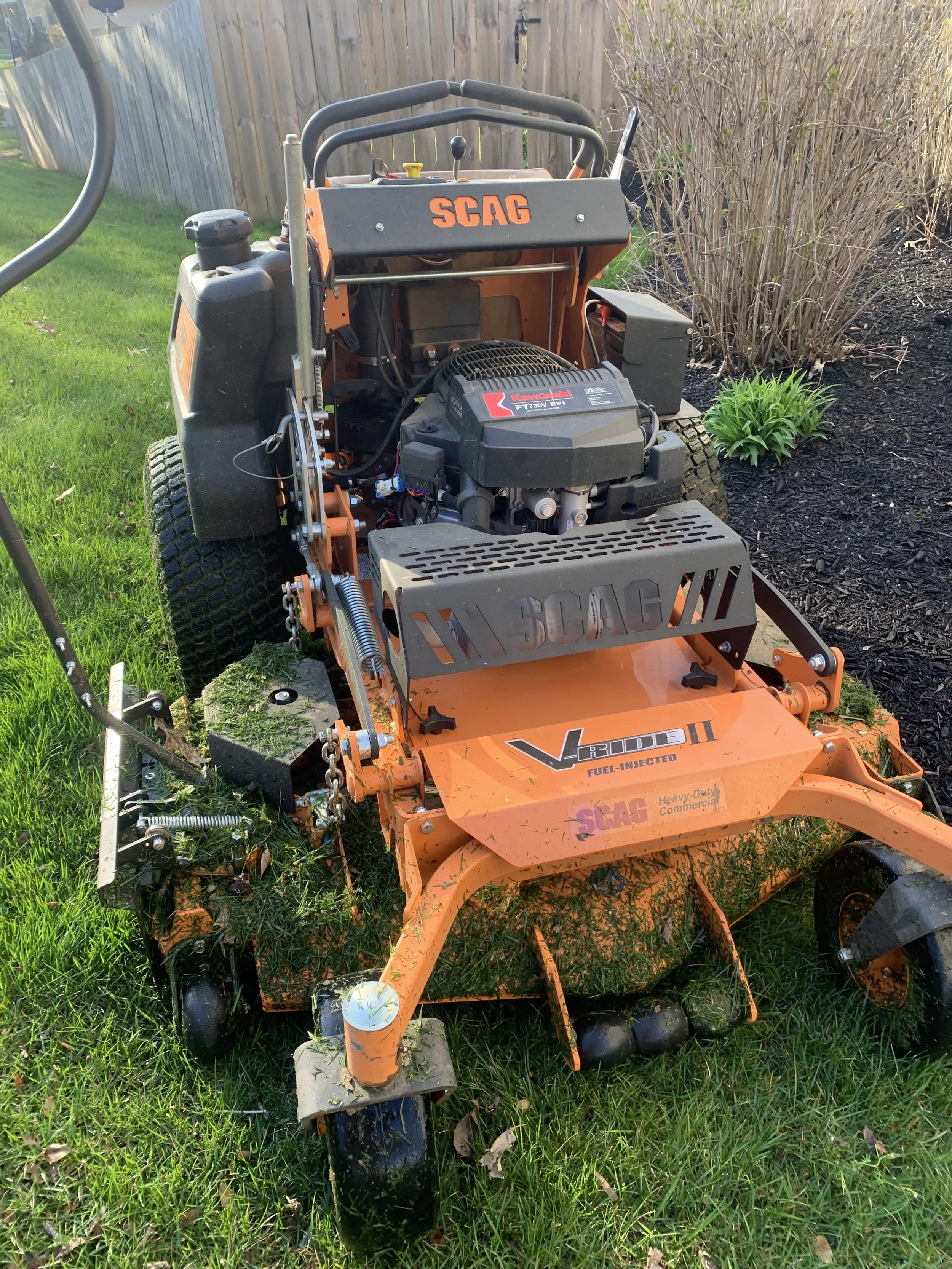 An orange SCAG V-Ride II commercial lawn mower parked on grass near a garden bed with shrubs.