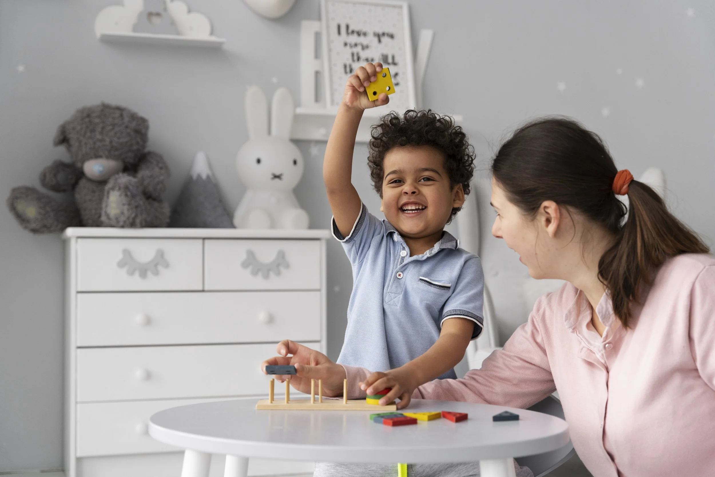 A young boy playing a game with a woman, smiling and holding a yellow game piece in a bright, decorated room.