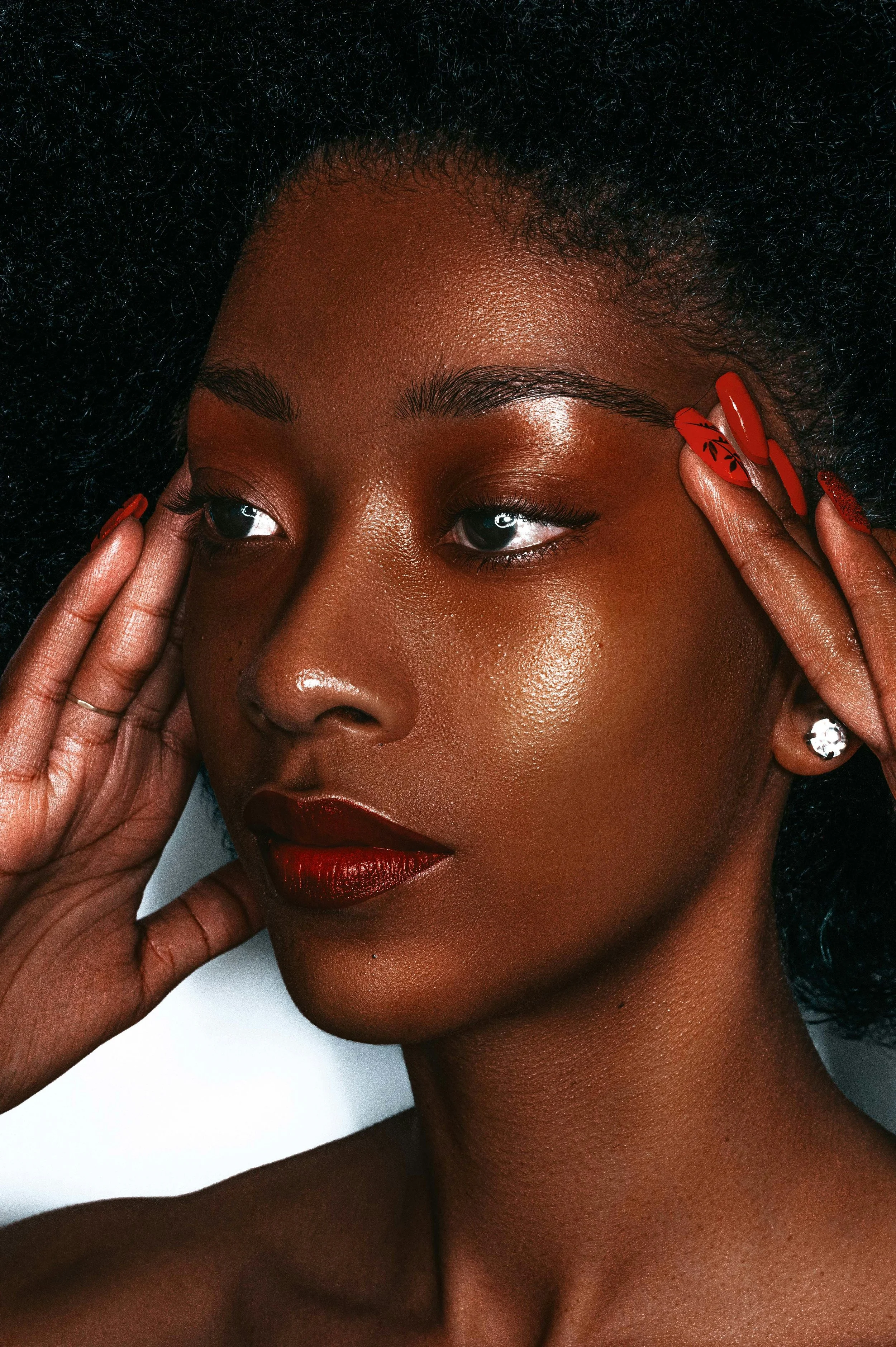 Close-up of an African-American woman's face with natural hair, bold red lipstick, red nails, and a large diamond earring, touching her temples.