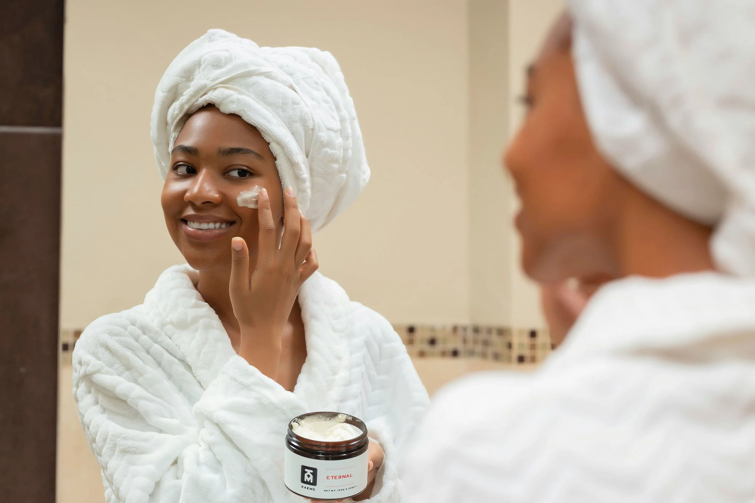 Young woman with a towel wrapped around her head applying facial cream while smiling and looking at herself in the mirror.