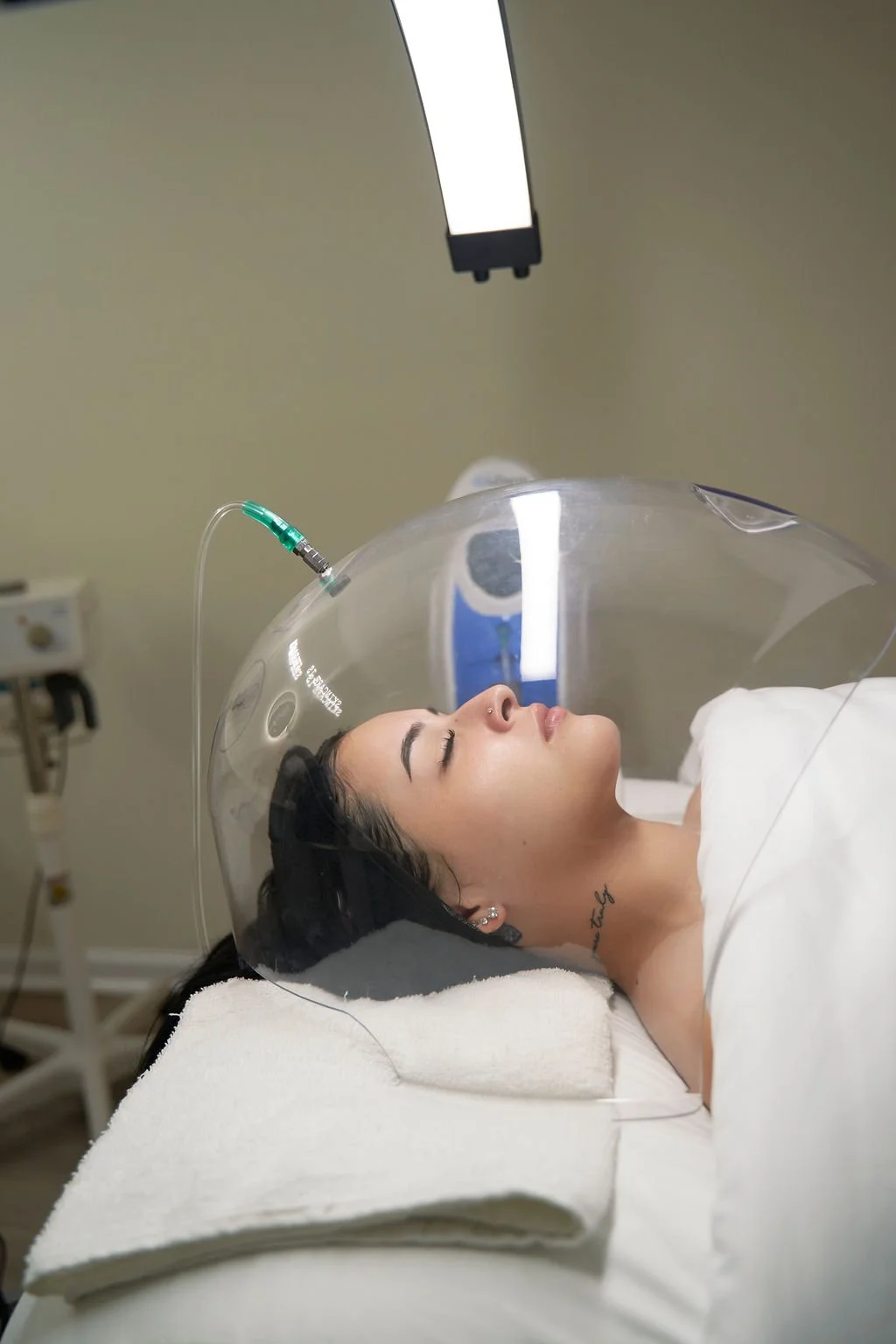 A woman lying in a medical chamber with a transparent dome over her head, connected to a medical device, in a clinical setting.