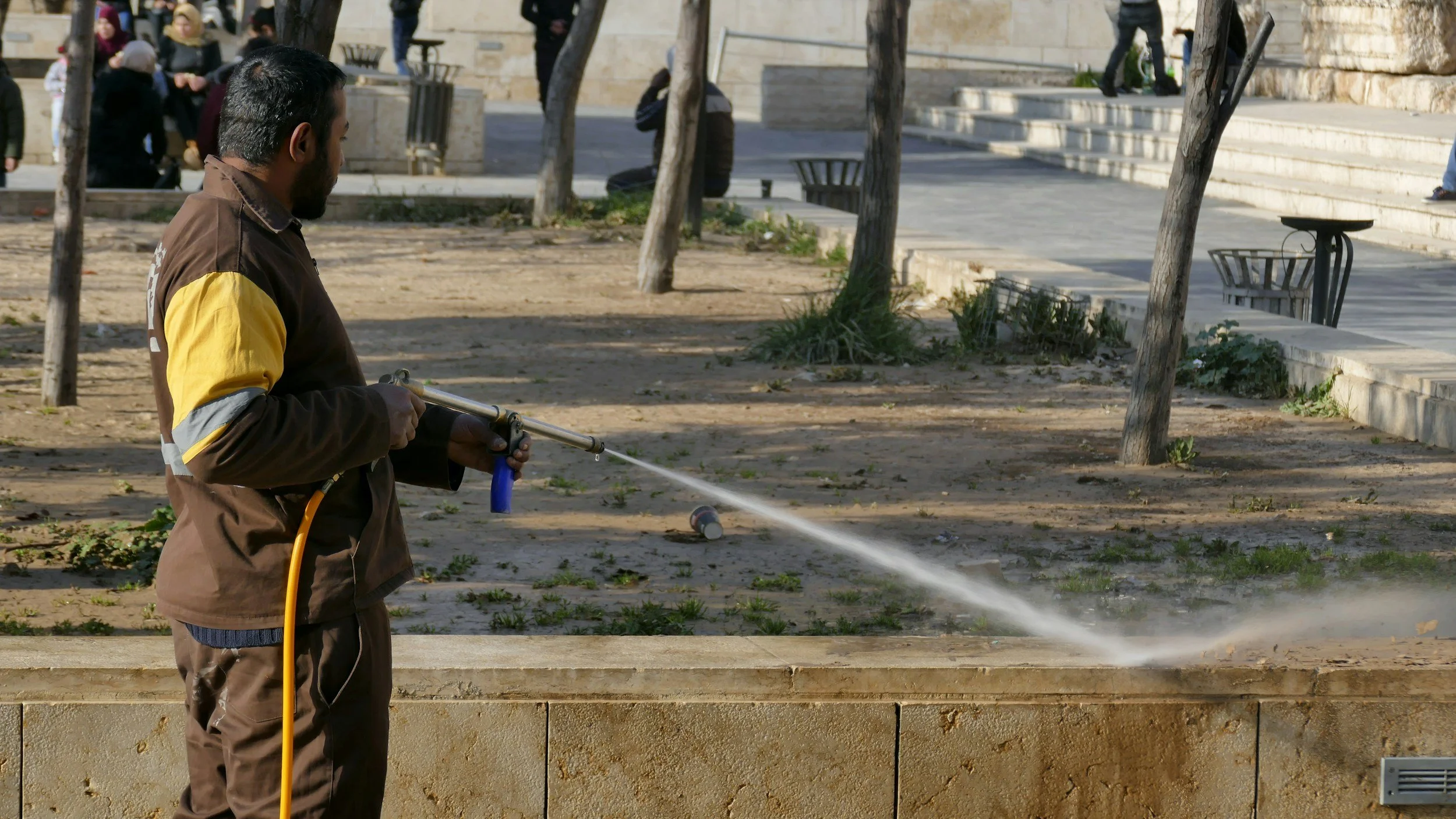 A man in a brown and yellow jacket is using a hose connected to a water source to spray water onto a stone ledge in a park, with trees, benches, and steps in the background.