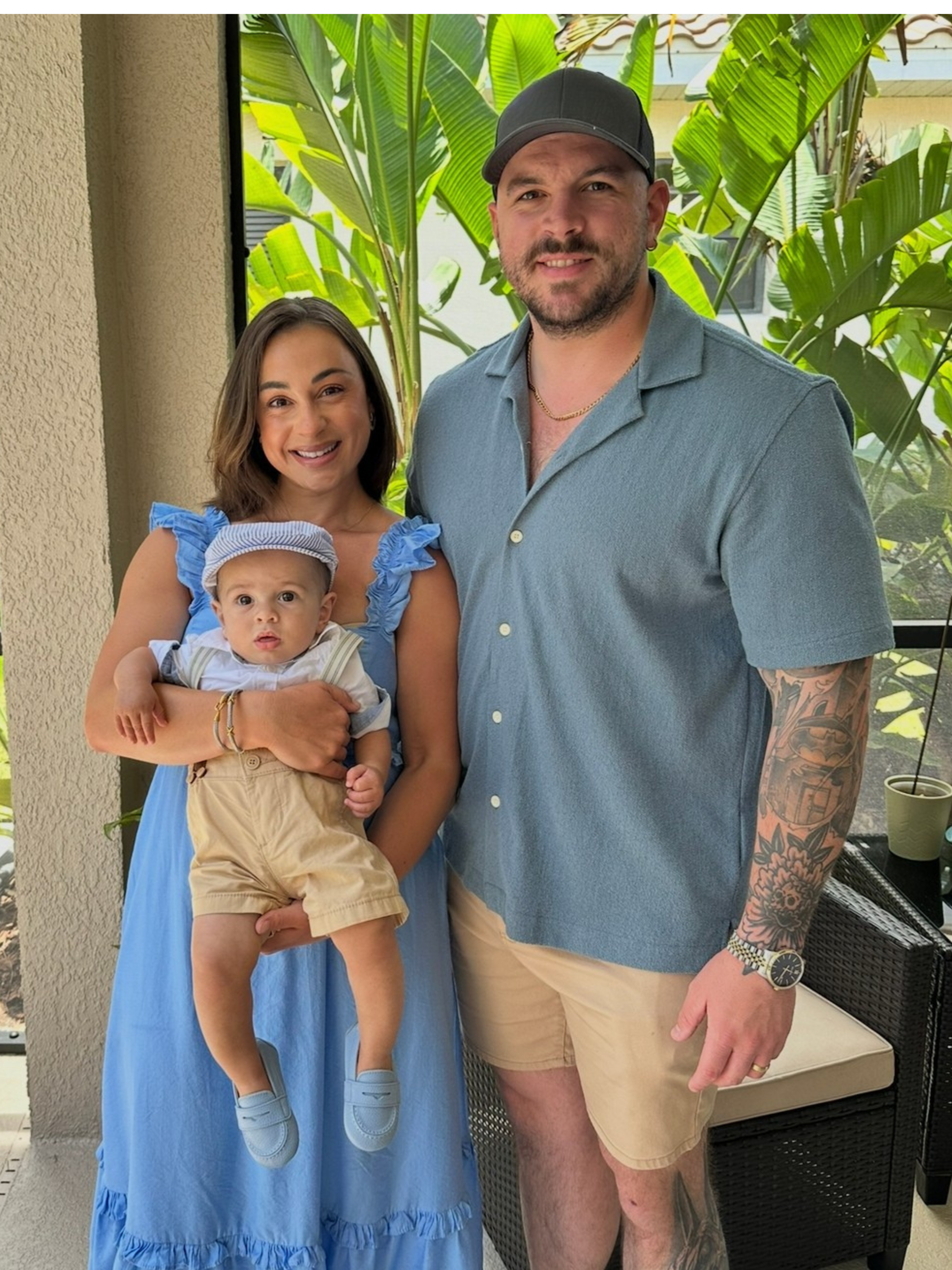 Family photo of a woman holding a baby, and a man standing beside them, with tropical plants in the background.