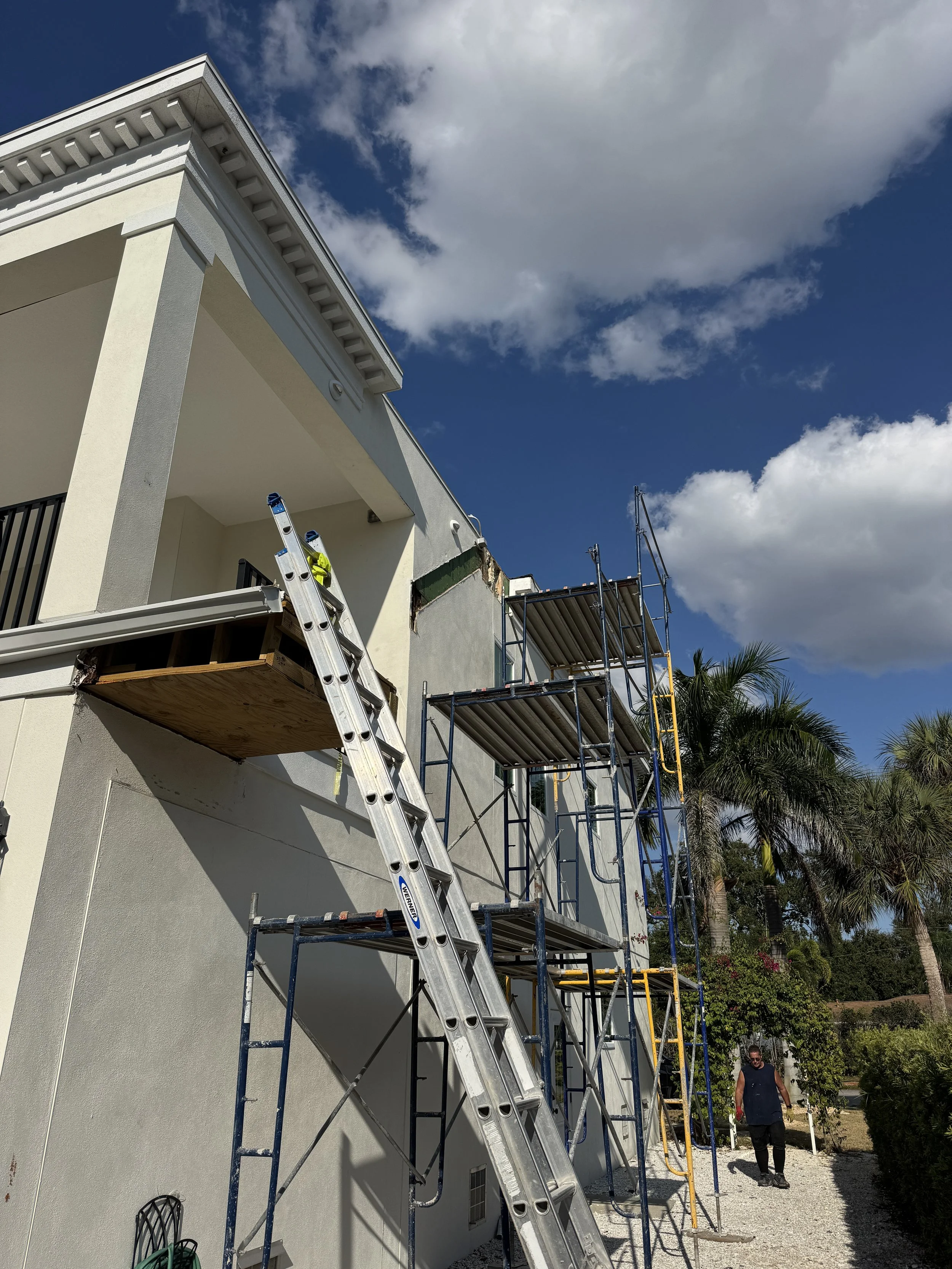 Construction workers working on the exterior of a building with scaffolding and a ladder, with clear sky and palm trees in the background.