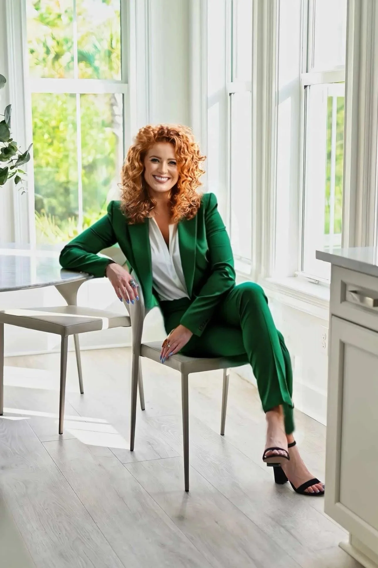 Gloria Black, award-winning luxury interior design consultant and creative strategist based in Naples Florida — professional portrait in emerald green suit seated in a bright light-filled studio with tropical greenery