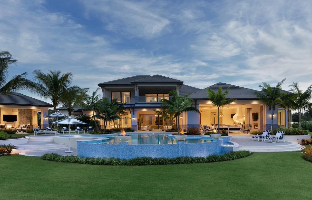 New England Charmer project by Gloria Black Design — luxury custom home exterior at twilight with illuminated facade, resort pool, and tropical landscaping, Naples Florida residential interior design