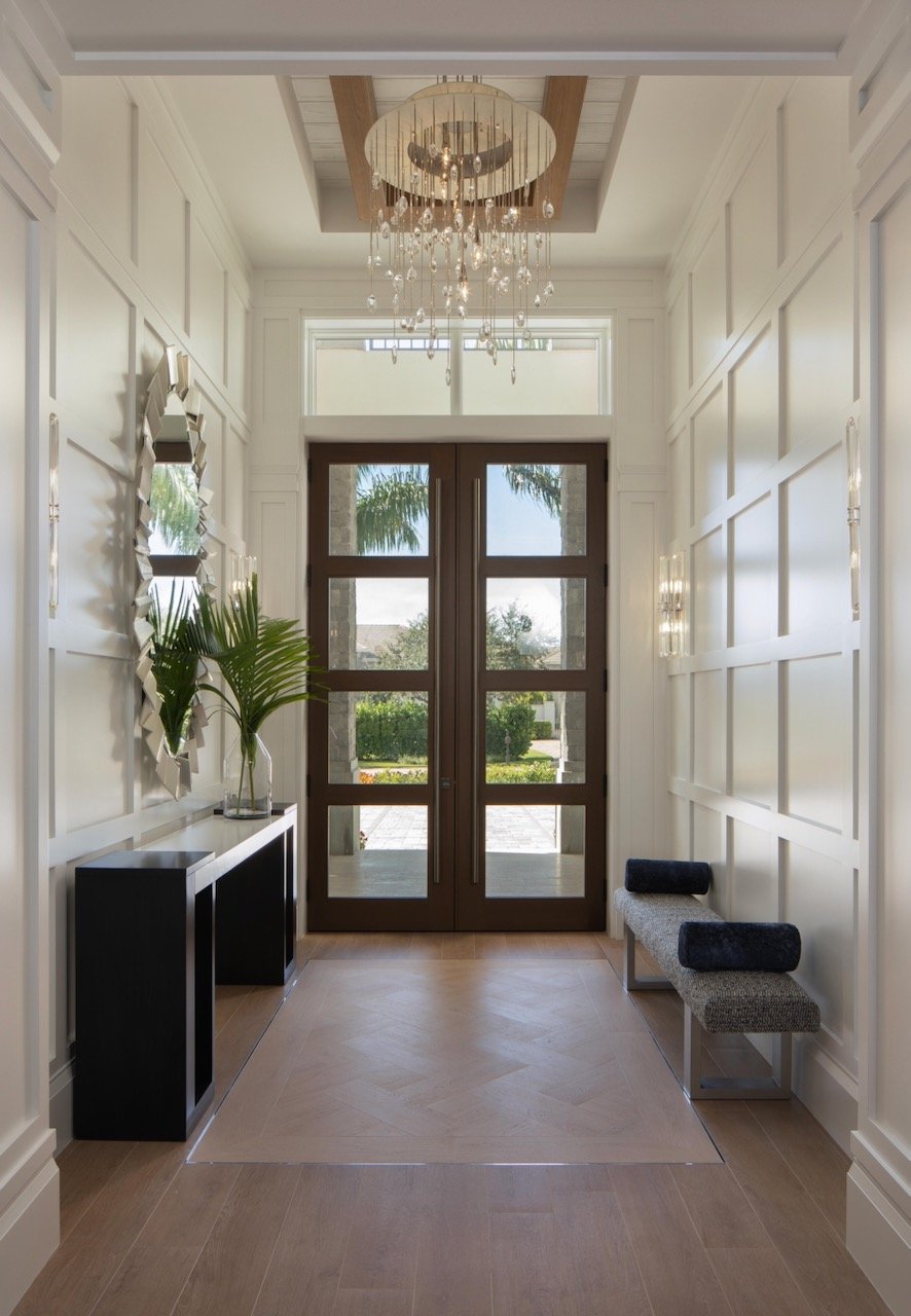 Luxury residential foyer with tall paneled walls, dark wood entry door and palm framing — Gloria Black Design Naples Florida