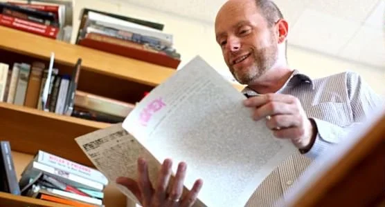 Padraic Kenney reading a newspaper in a room with bookshelves.