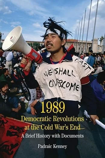 Protester holding a megaphone and wearing a sign that says 'WE SHALL OVERCOME' during a demonstration, with a crowd in the background, and book cover for '1989 Democratic Revolutions at the Cold War's End' by Padraic Kenney.