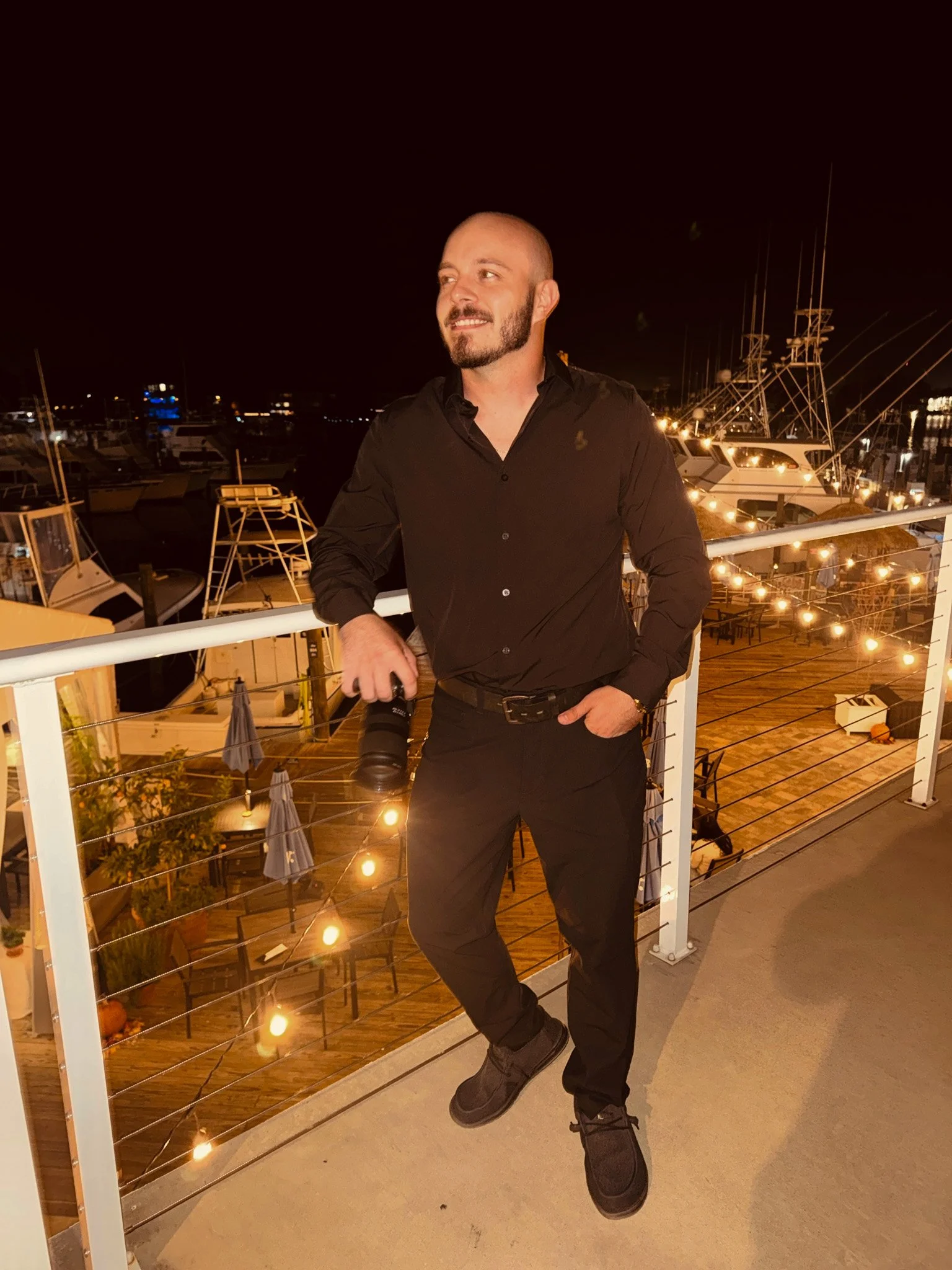 A man in a black shirt and black pants standing on a balcony at night, holding a camera, with boats and string lights in the background.