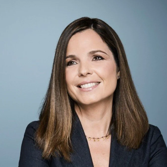 A woman with shoulder-length brown hair smiling, wearing a dark blazer and a gold necklace, against a plain blue background.