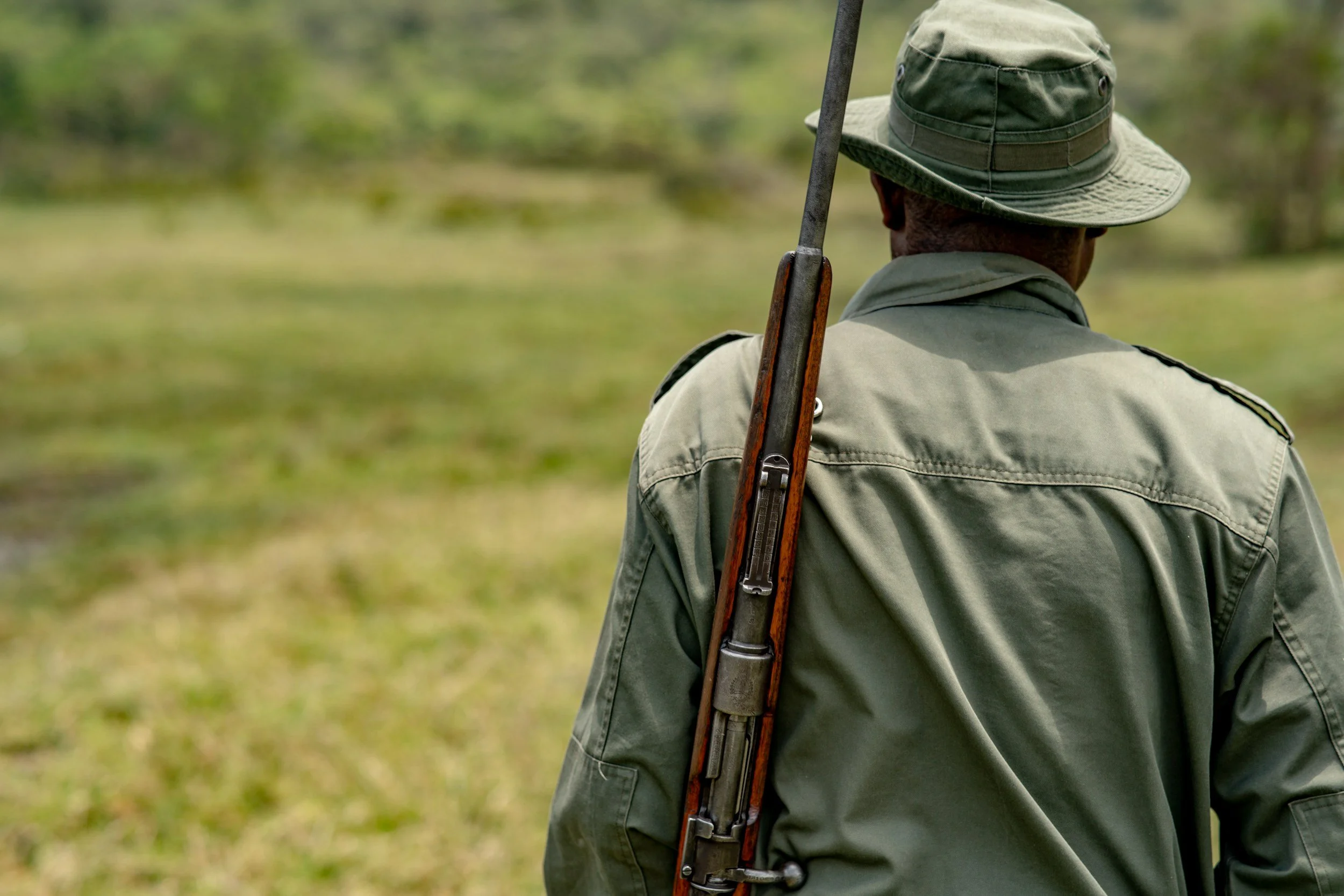 Back view of a person wearing a green hat and military-style uniform holding a rifle over their shoulder, outdoors on a grassy field.