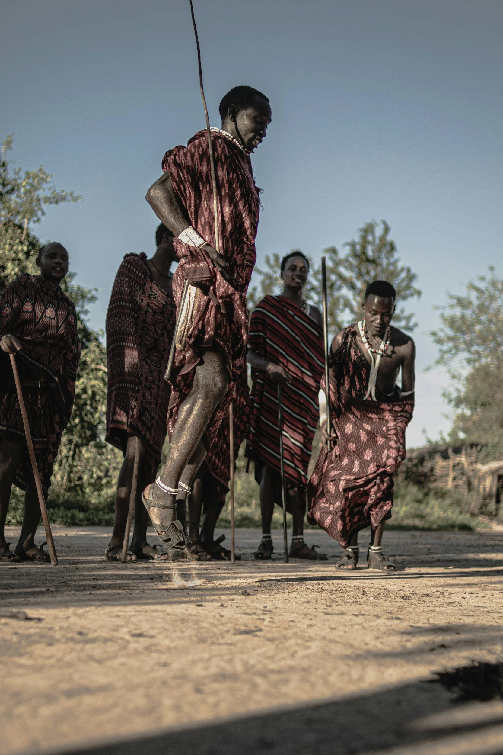 Group of Maasai men in traditional attire standing outdoors, some holding sticks, with trees and a clear sky in the background.