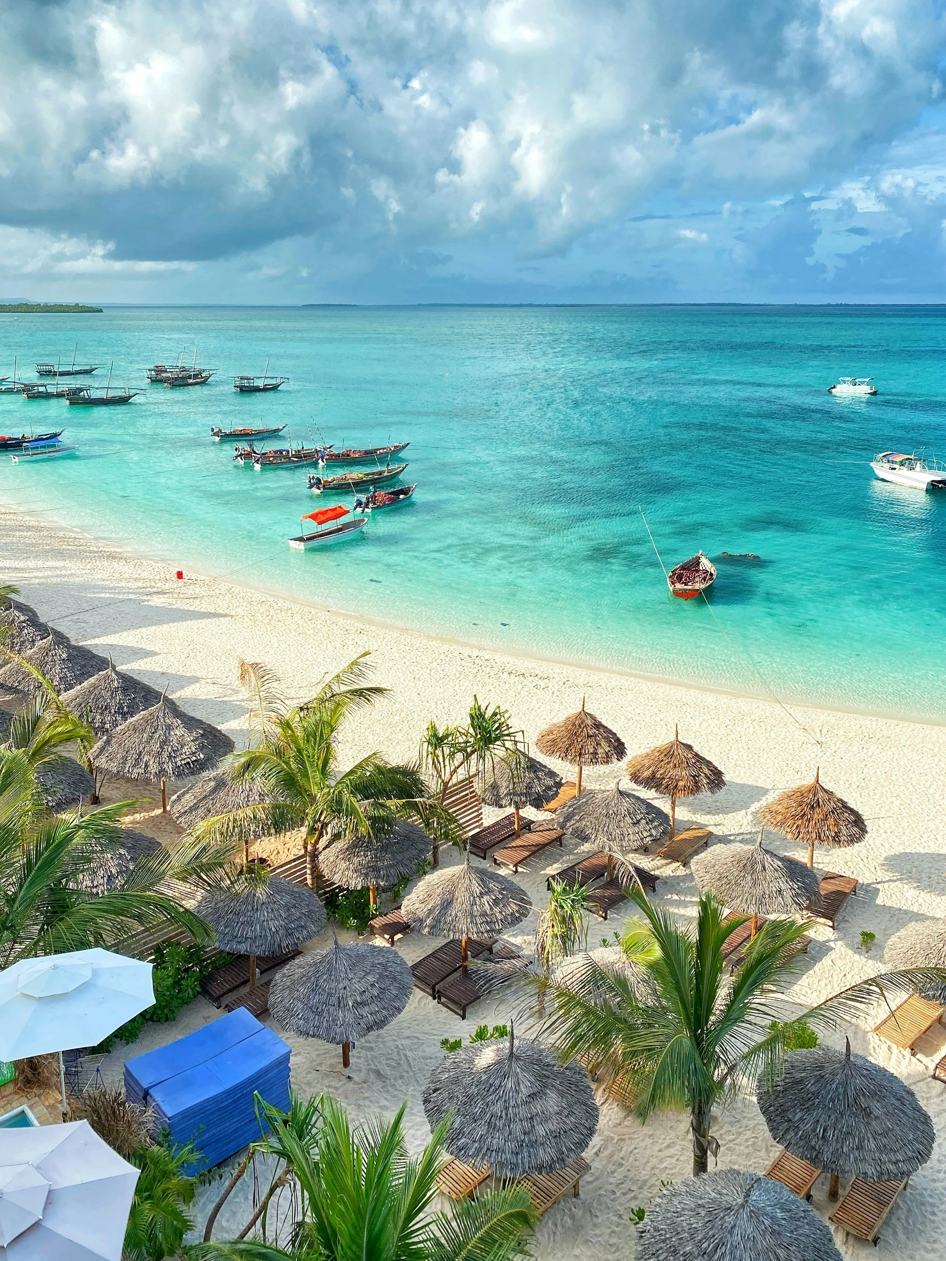A tropical beach with thatched umbrellas, lounge chairs, palm trees, and turquoise water with boats anchored offshore under a partly cloudy sky.