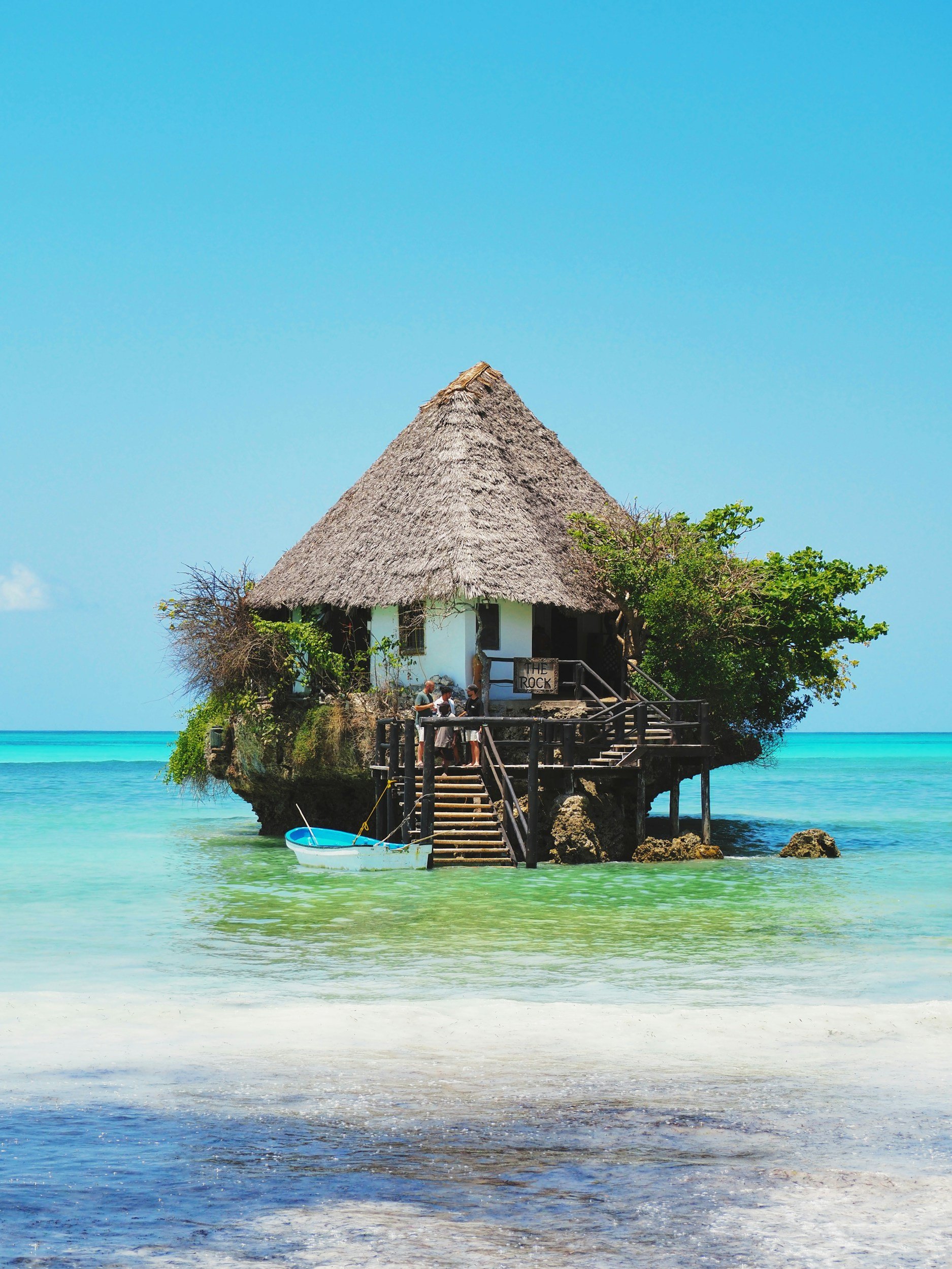 A thatched roof hut on stilts over turquoise water with a small boat docked nearby, under a clear blue sky.