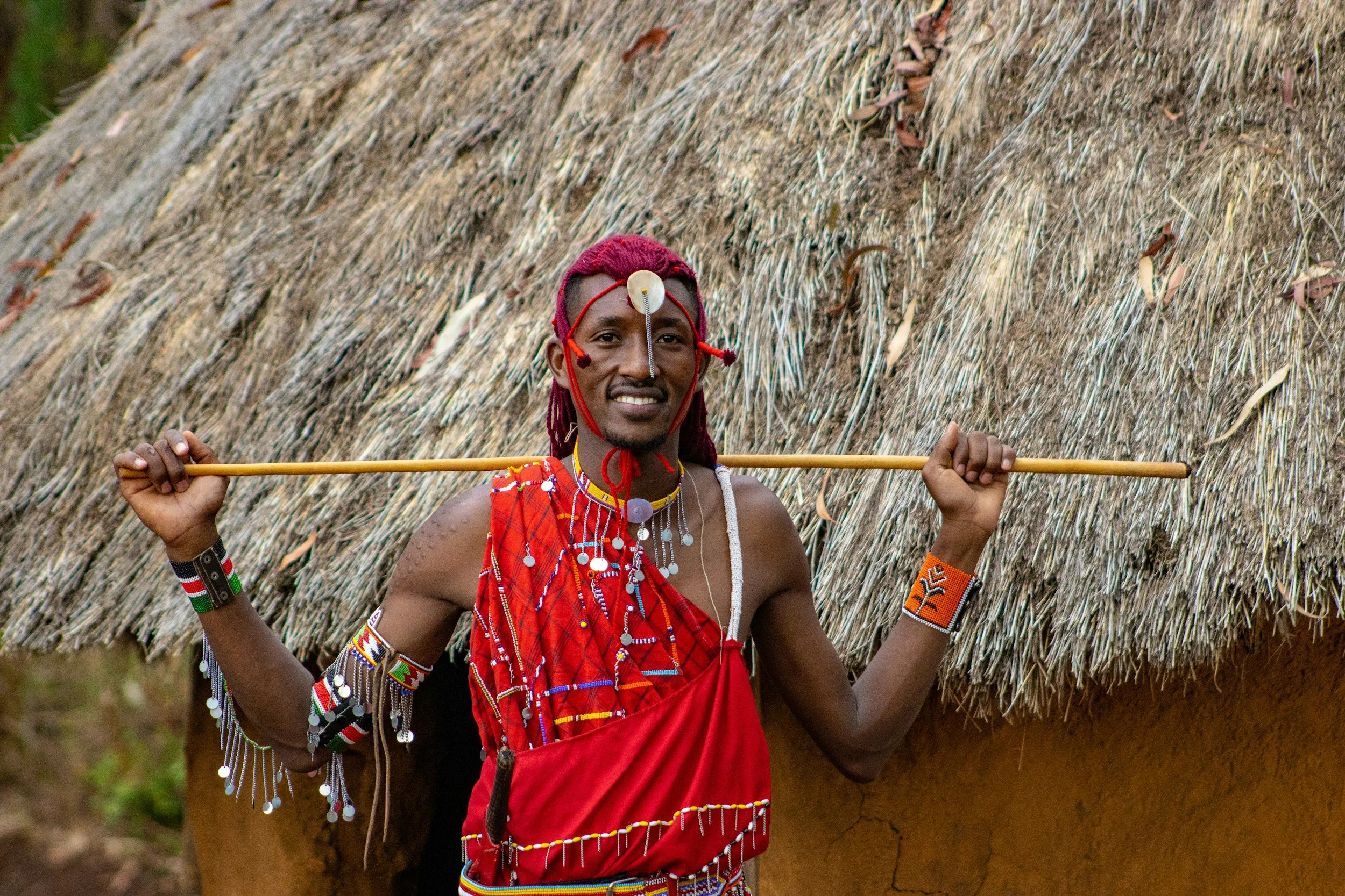A man dressed in traditional Maasai attire, wearing beaded jewelry, a red shuka, and holding a spear, standing in front of a thatched hut.