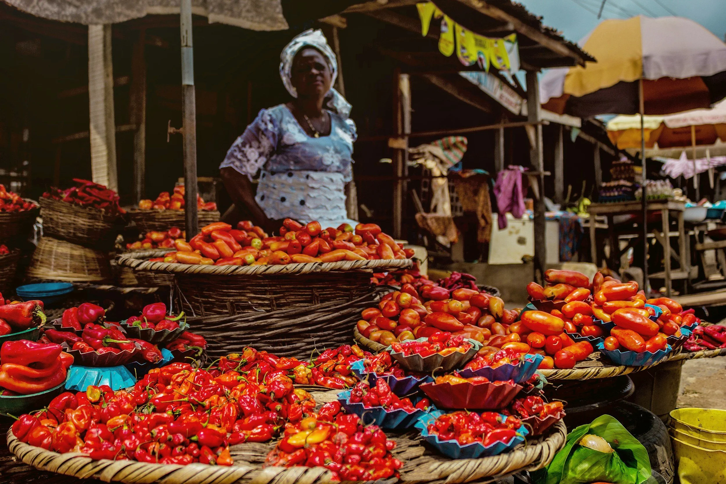 A woman standing at a market stall filled with an array of red and orange hot peppers and tomatoes in woven baskets.