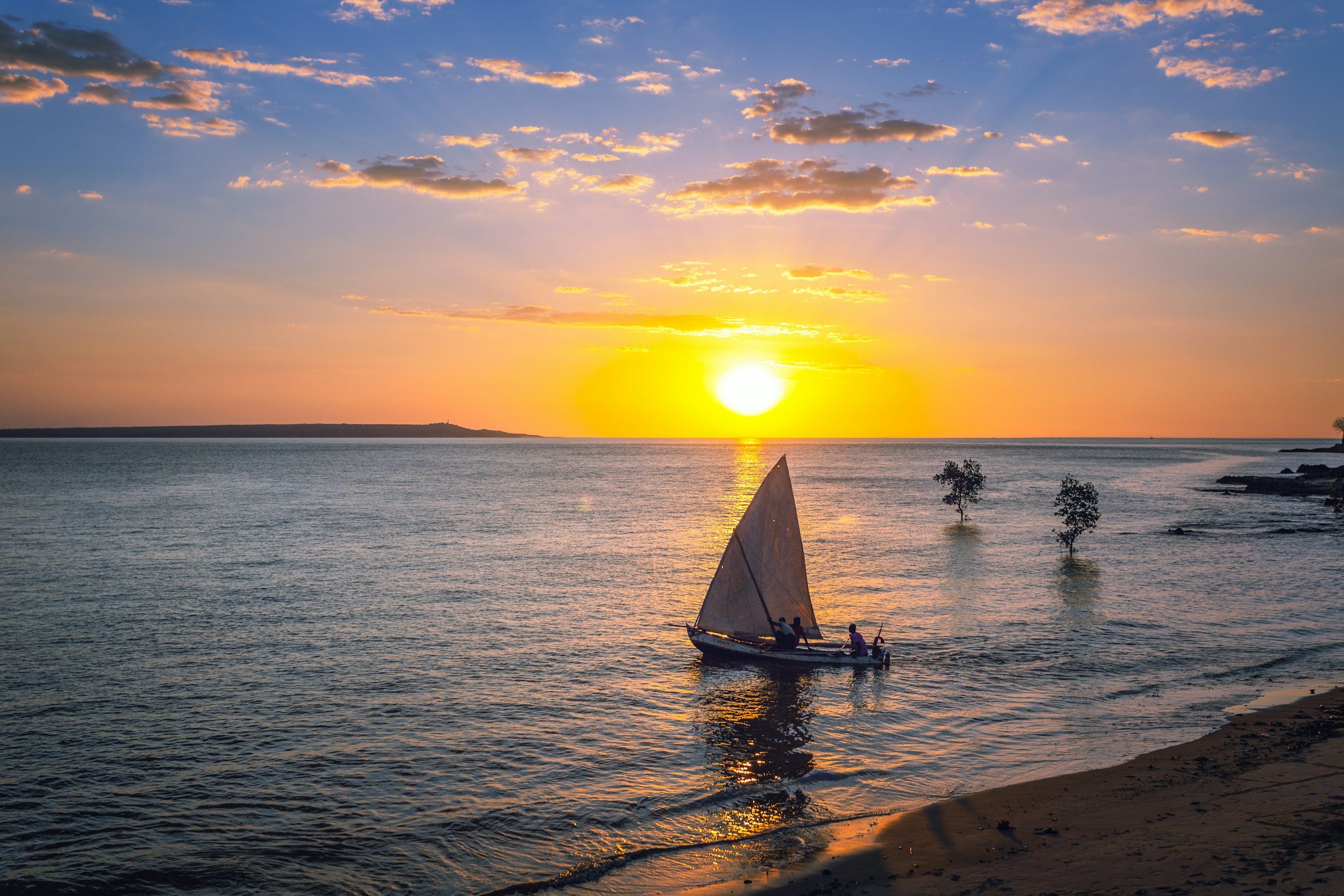 A sailboat with three people on the water during a sunset, with clouds in the sky and trees along the shoreline.