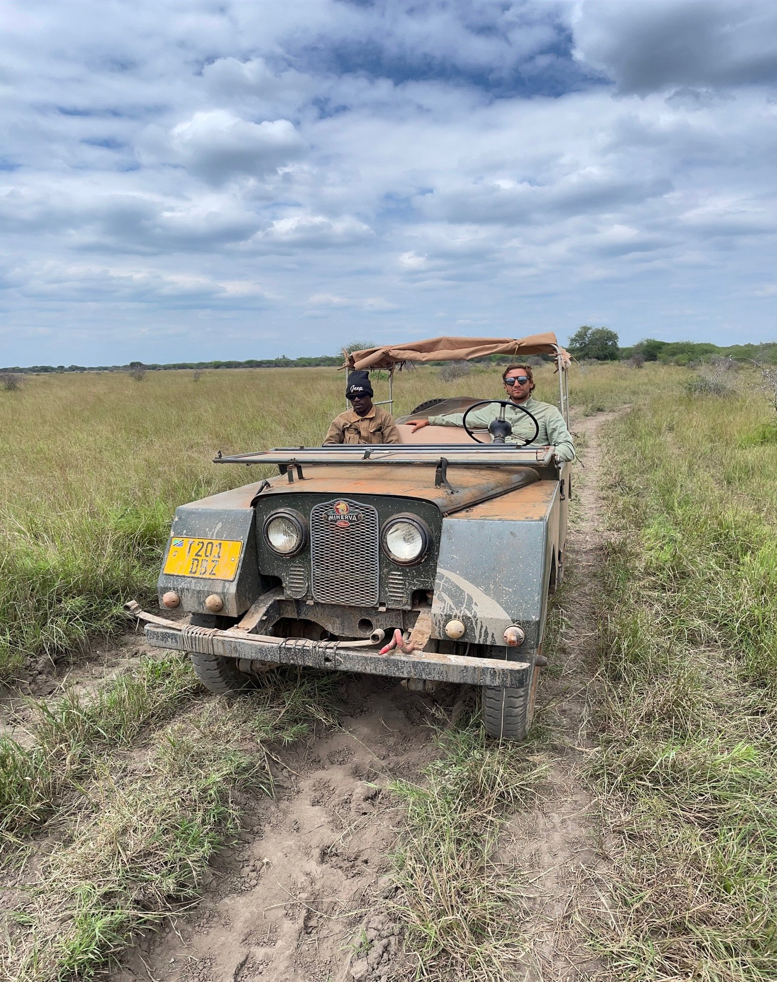 Two men riding in an open-top beige off-road vehicle in a grassy plain under a cloudy sky, with dirt track visible.