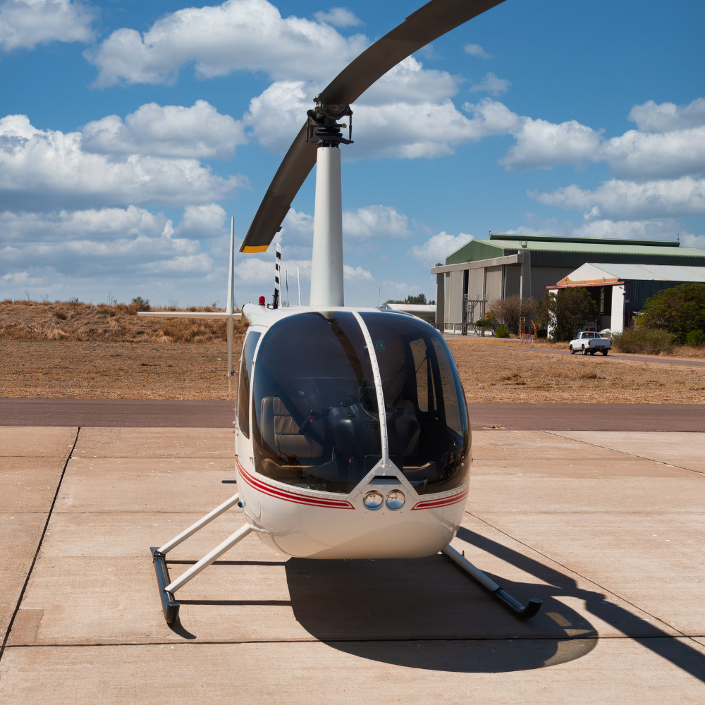 A small white helicopter with a bubble-shaped cockpit parked on a tarmac under a partly cloudy sky.