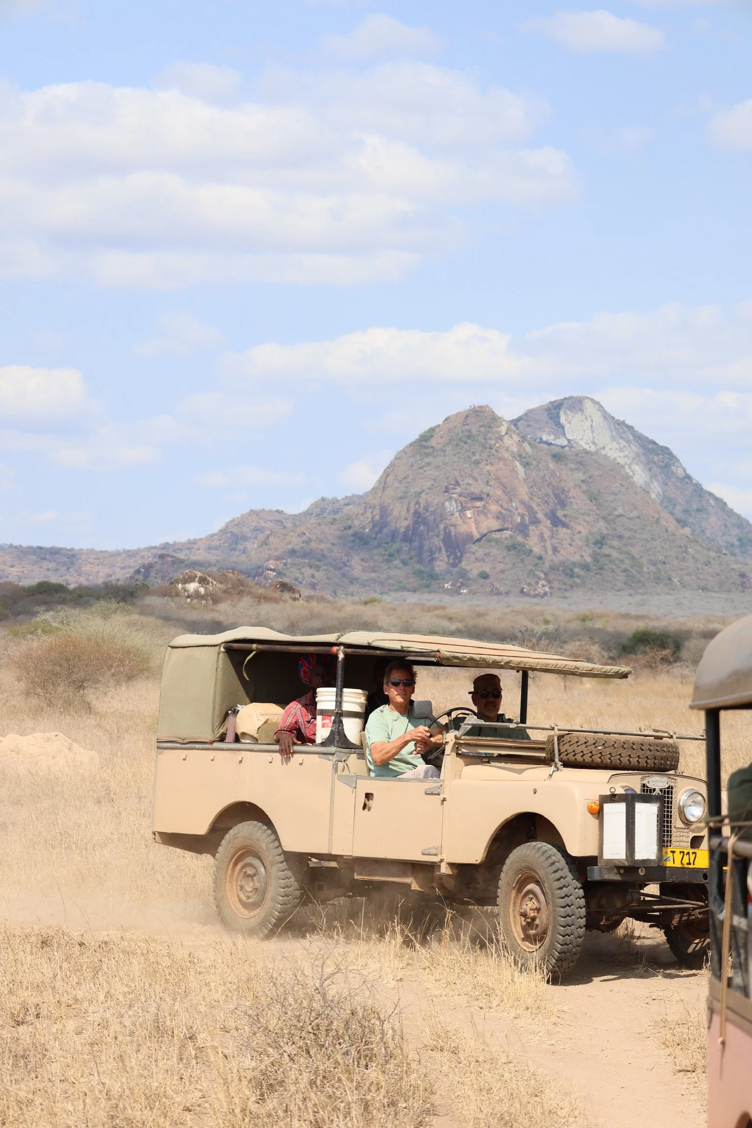People riding in an off-road vehicle in a dry, hilly landscape with a mountain in the background and a partly cloudy sky.