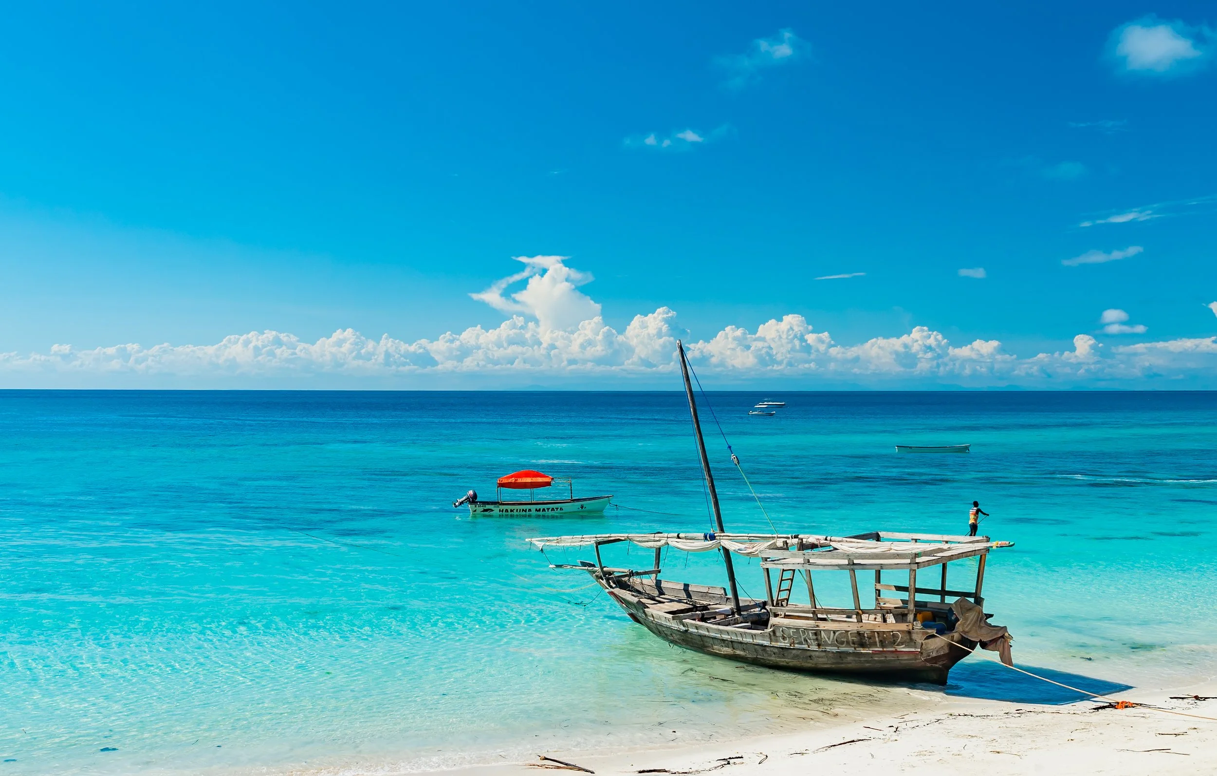 Clear turquoise ocean with boats and a person paddleboarding near a sandy beach, under a blue sky with scattered clouds.