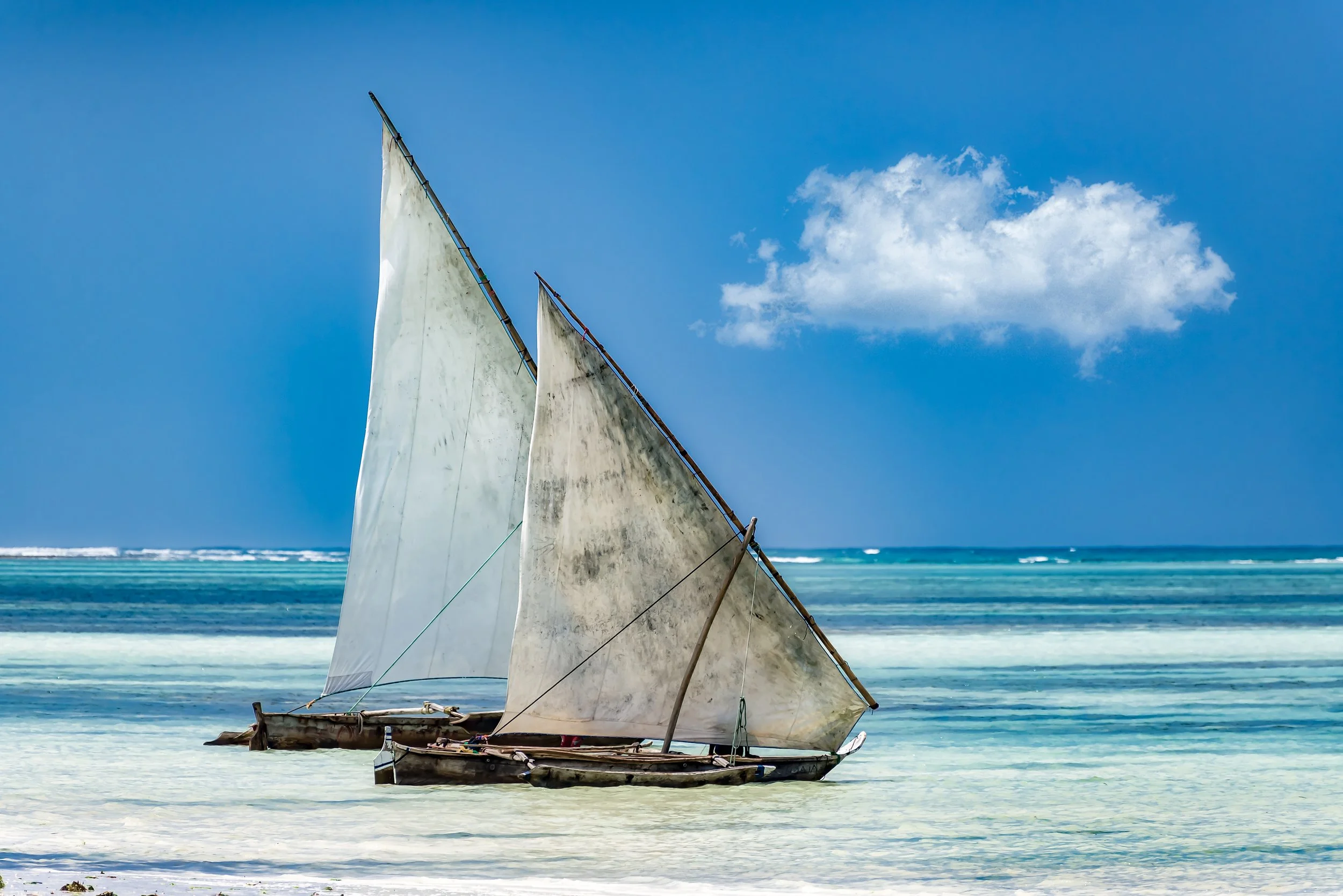An old wooden sailboat with tattered sails resting on a sandy beach with clear blue ocean and sky in the background.