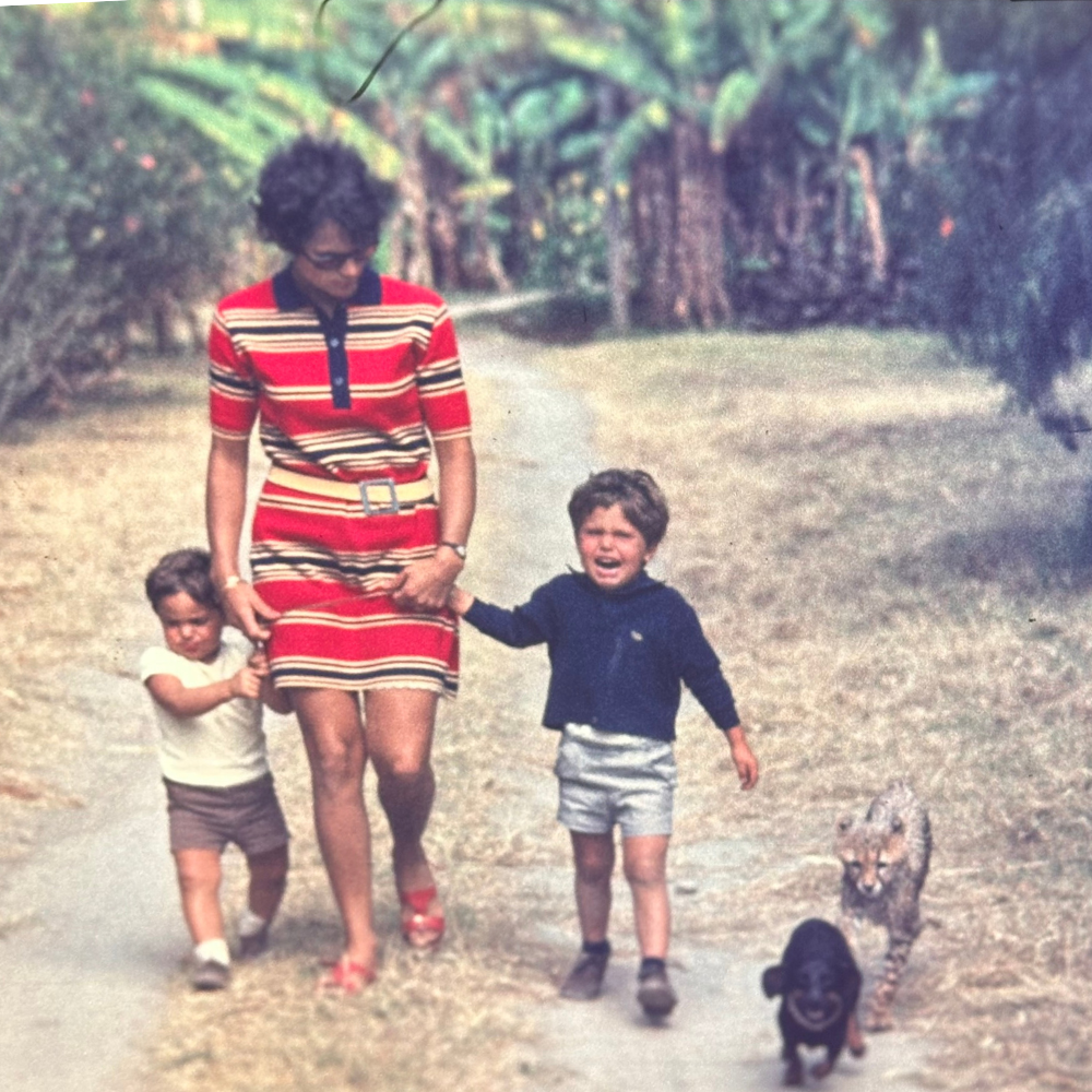 A woman walking on a dirt path with two young children and two animals, a small black dog and a baby cheetah, in a wooded area.