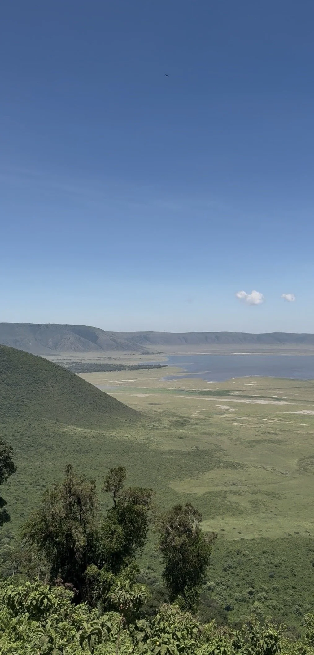 Scenic view of green rolling hills leading to a valley with water, situated in front of distant mountains under a clear blue sky with a few small clouds.