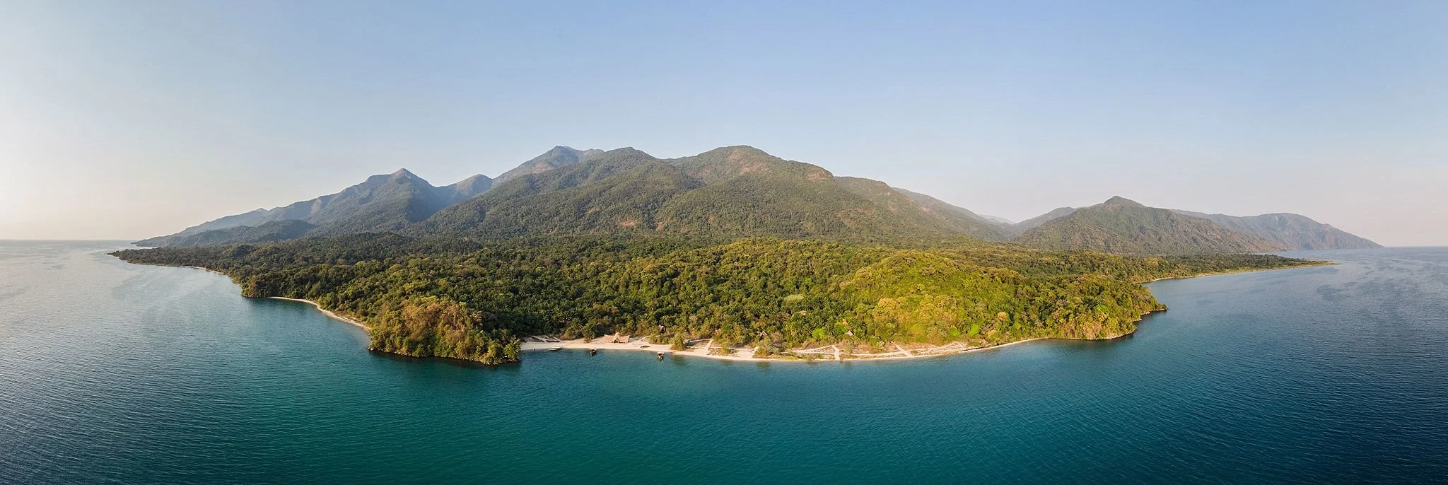A panoramic aerial view of a lush green island surrounded by calm blue waters with mountains in the background under a clear sky.