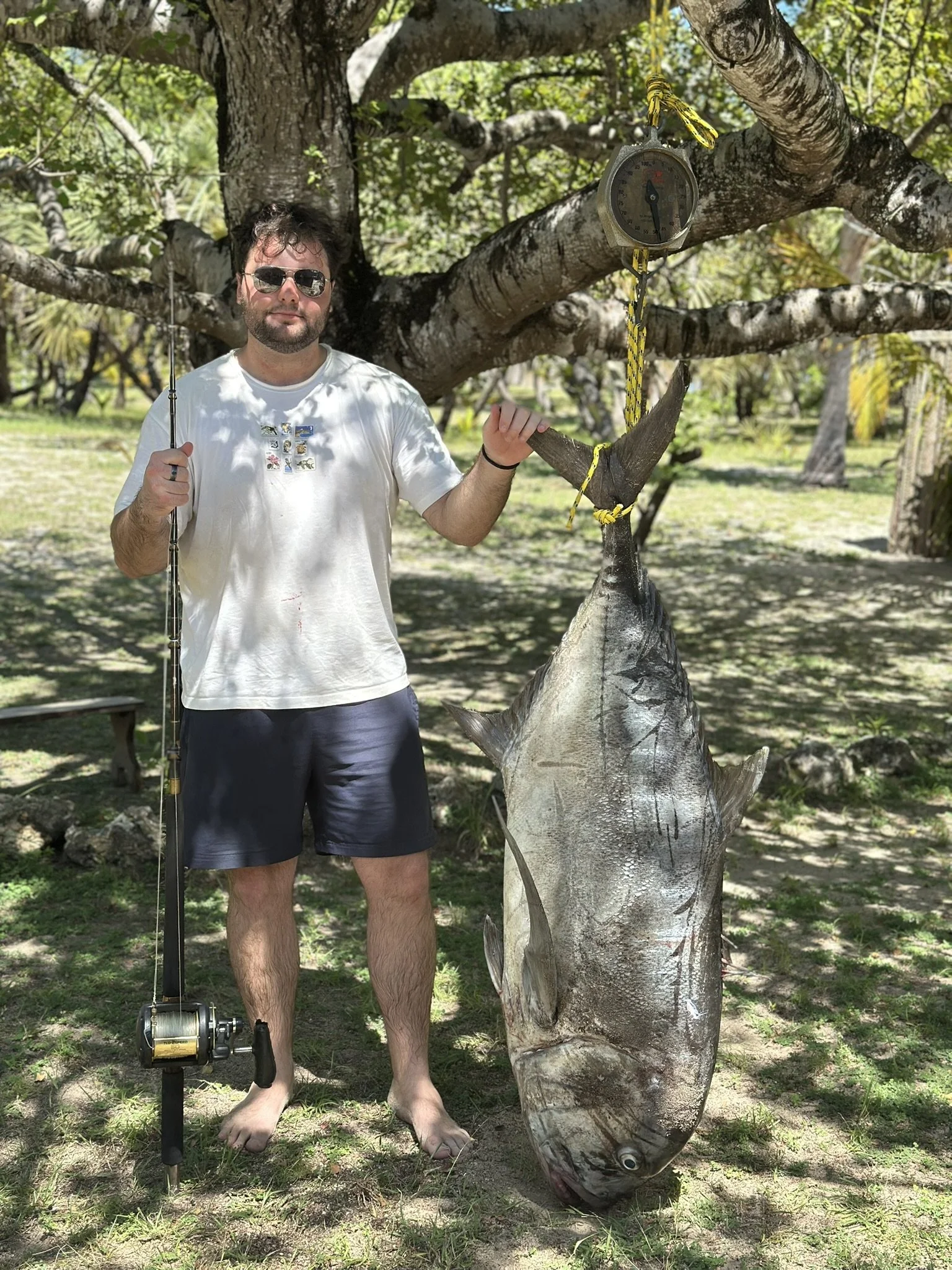 A man with sunglasses, white t-shirt, shorts, and barefoot stands next to a large fish caught from the water, hanging on a tree. The man holds a fishing rod with one hand and touches the fish with the other. The scene is outdoors with trees and grass, and a measuring scale hangs from the tree.