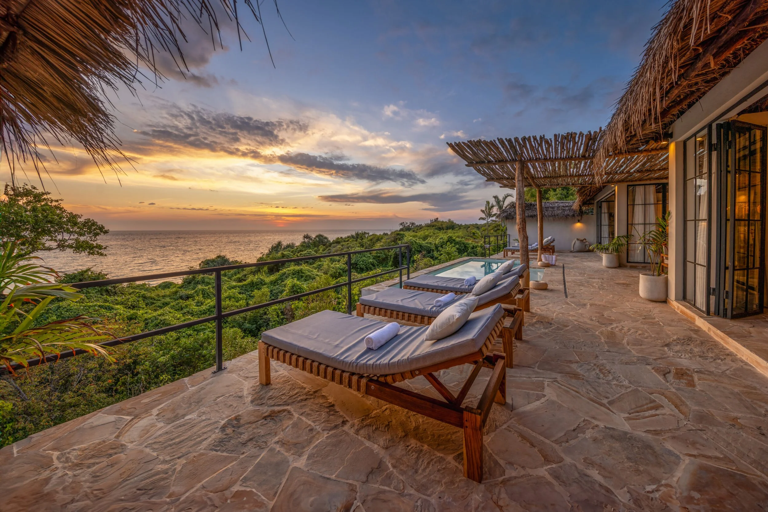 Balcony with lounge chairs overlooking a forest and ocean sunset, with thatched roof shades and glass doors.