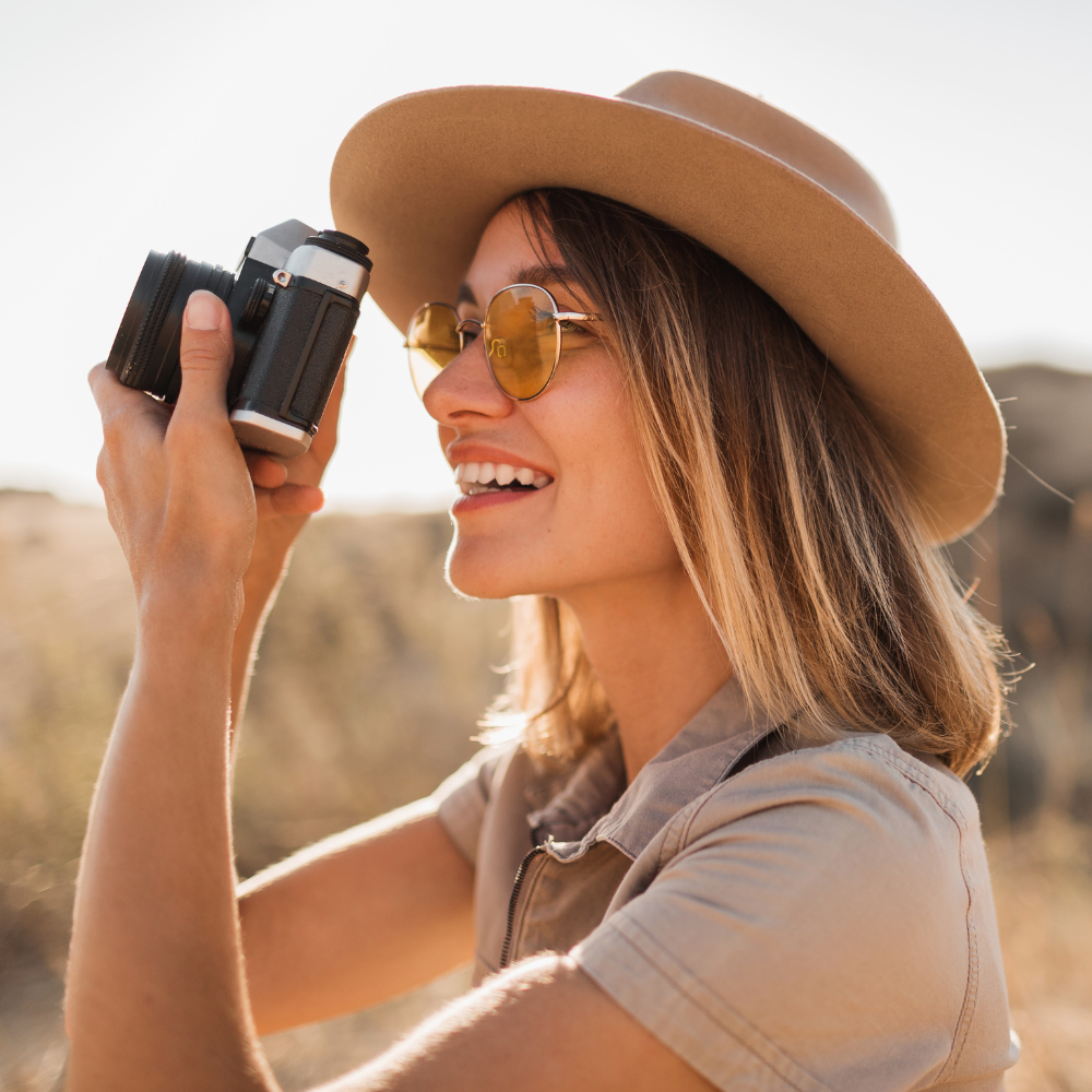 A woman with a tan hat, sunglasses, and a beige shirt holding a vintage camera near her face outdoors during daytime.