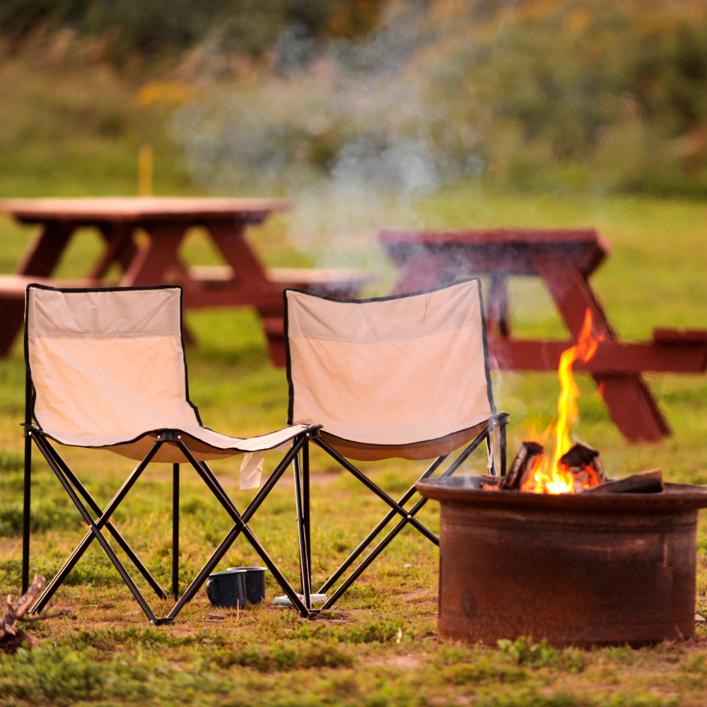 Two white camping chairs beside a small campfire in a grassy area with discolored picnic tables in the background
