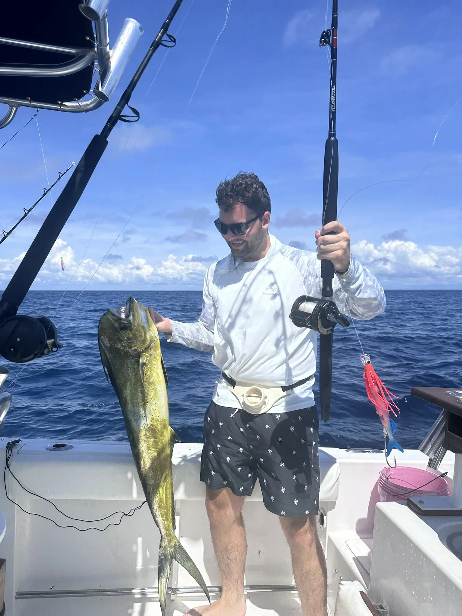A man holding a large fish on a boat with fishing rods and equipment visible, under a clear blue sky over the ocean.