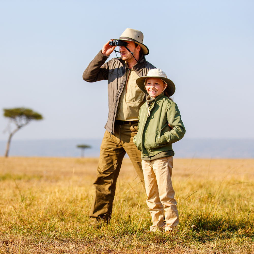 A man and a young girl in safari attire standing in a grassy field, with the man looking through binoculars and the girl smiling, wearing hats and jackets.