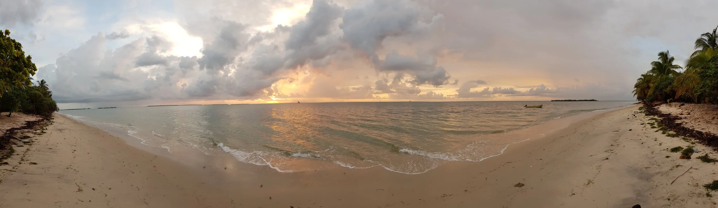 A panoramic view of a tropical beach at sunset with cloudy sky, calm ocean water, palm trees on the right, and a small boat floating near the shore.