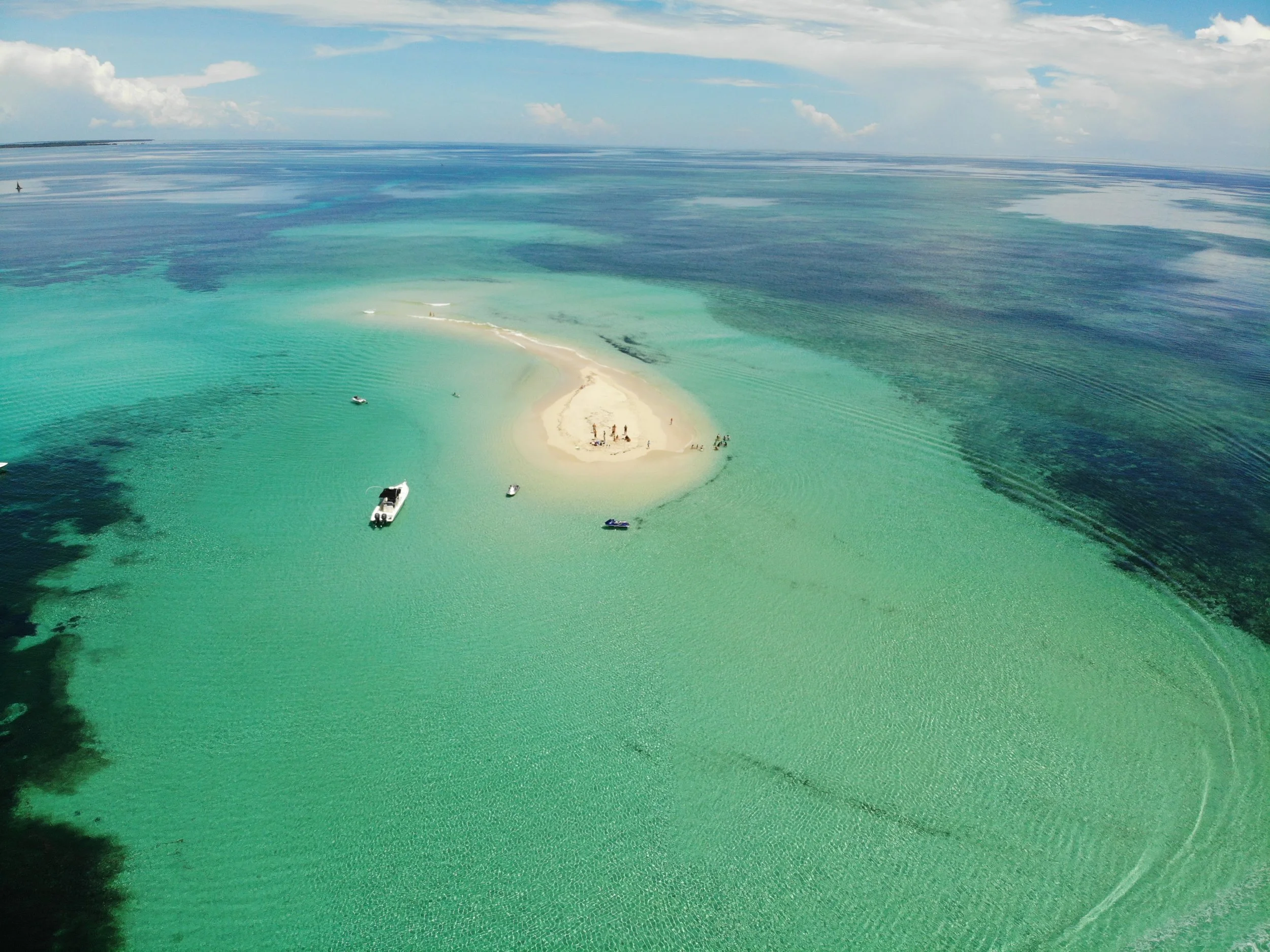 Aerial view of a small sandy island surrounded by clear turquoise ocean water, with boats floating nearby and a person walking on the island, under a partly cloudy sky.