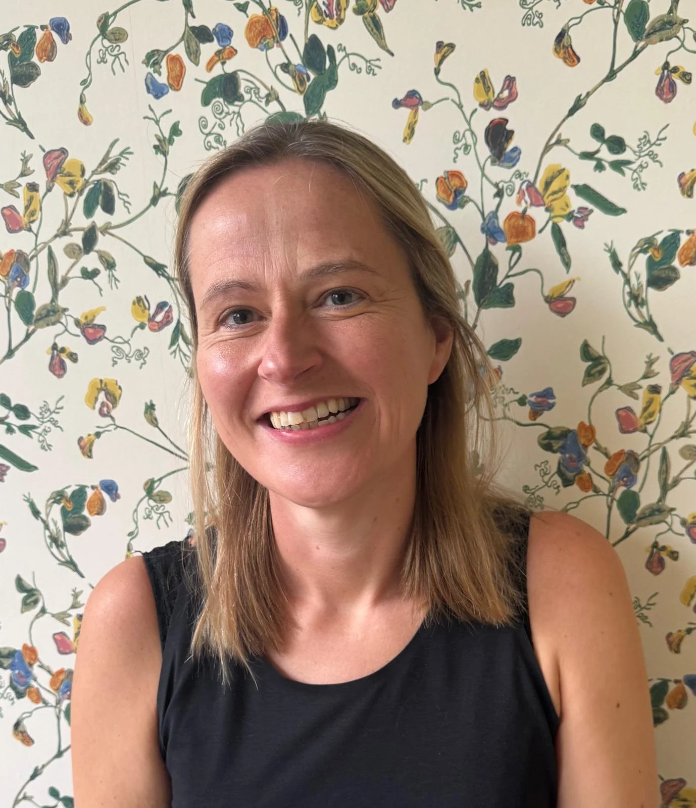 image of Clare smiling -   shoulder-length blonde hair wearing a black sleeveless top, standing in front of a decorated wall with a colorful floral pattern.
