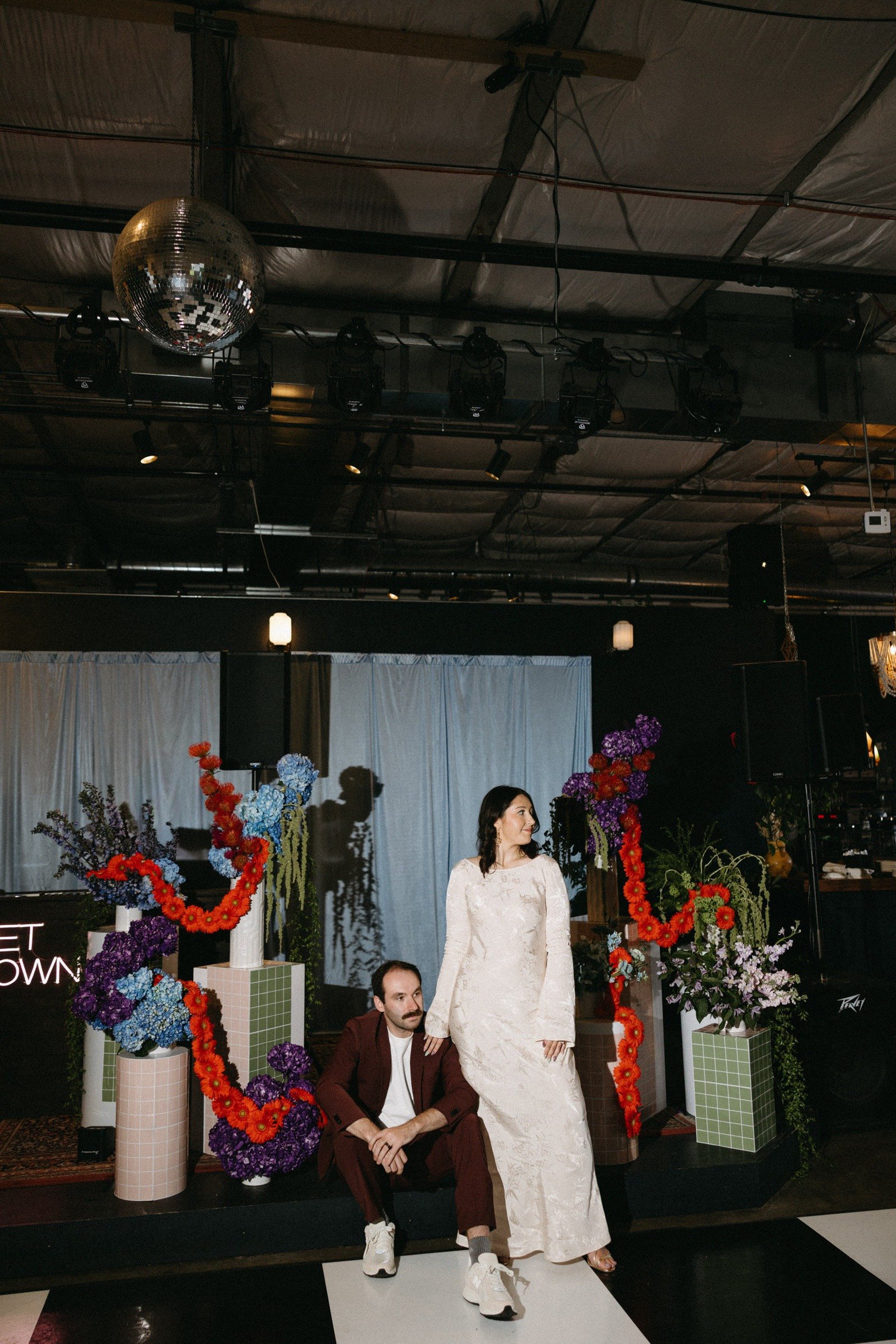A woman in an elegant wedding dress standing next to a man in casual maroon suit and white sneakers, sitting on a stage with colorful floral arrangements and geometric vases. The background features a white curtain and dim lighting, with a disco ball