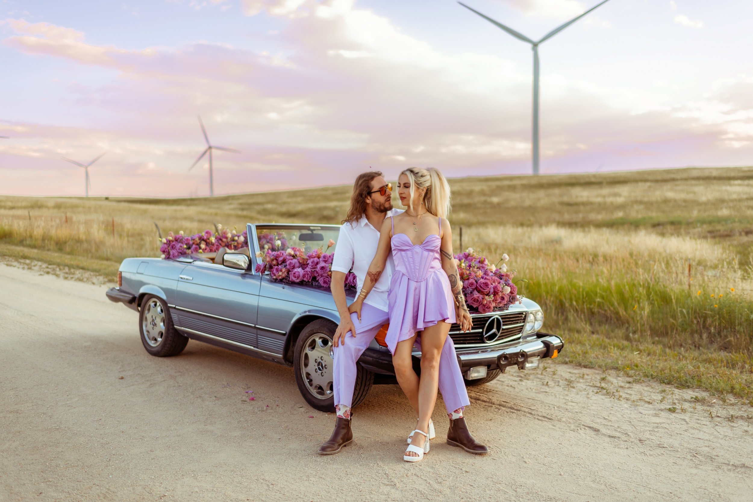 A man and woman leaning against a vintage Mercedes convertible decorated with pink and purple flowers on an open dirt road with wind turbines in the background during sunset.