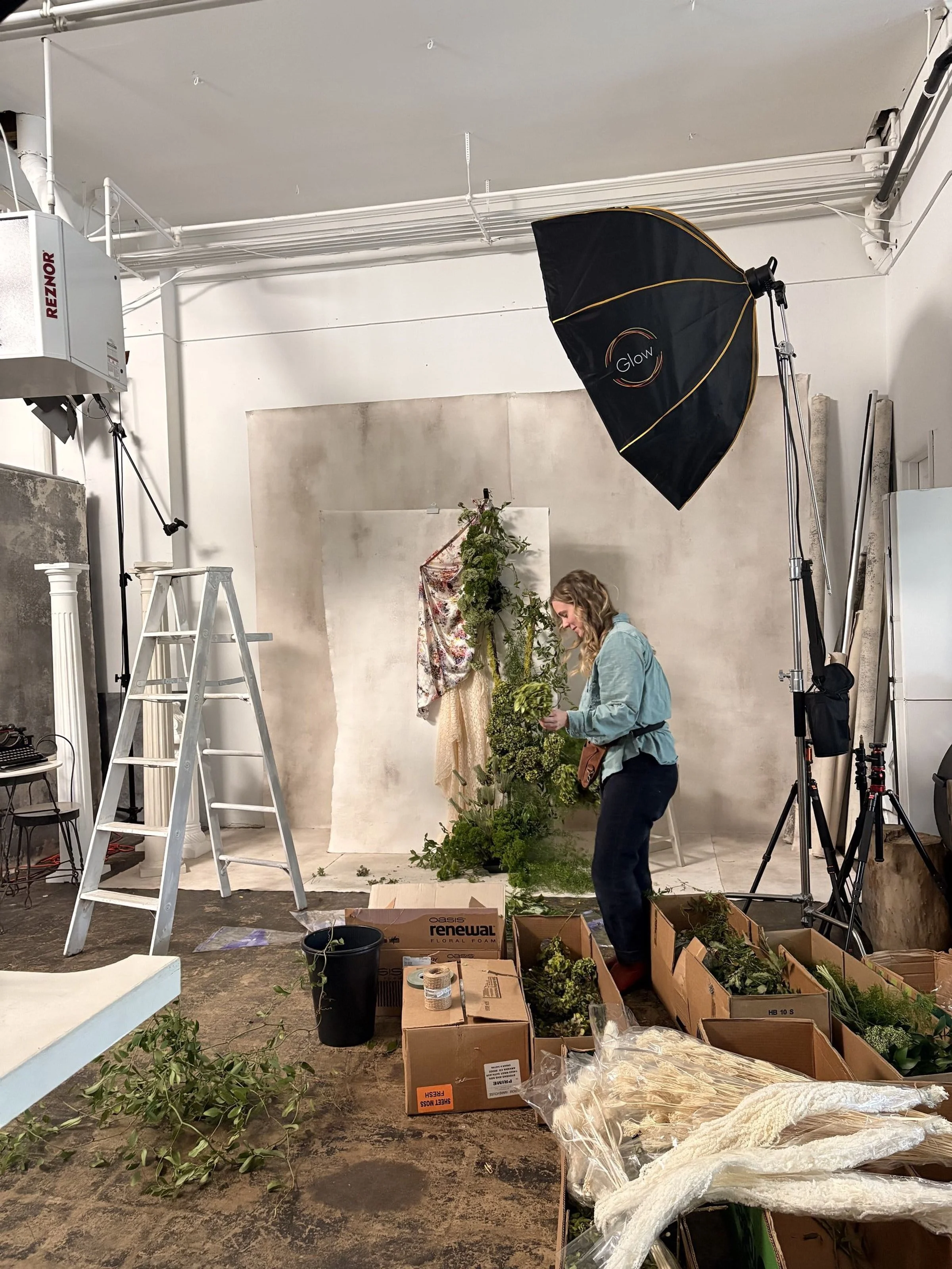 A woman is preparing floral arrangements in a photography studio with professional lighting equipment, ladders, and various boxes of greenery and flowers.