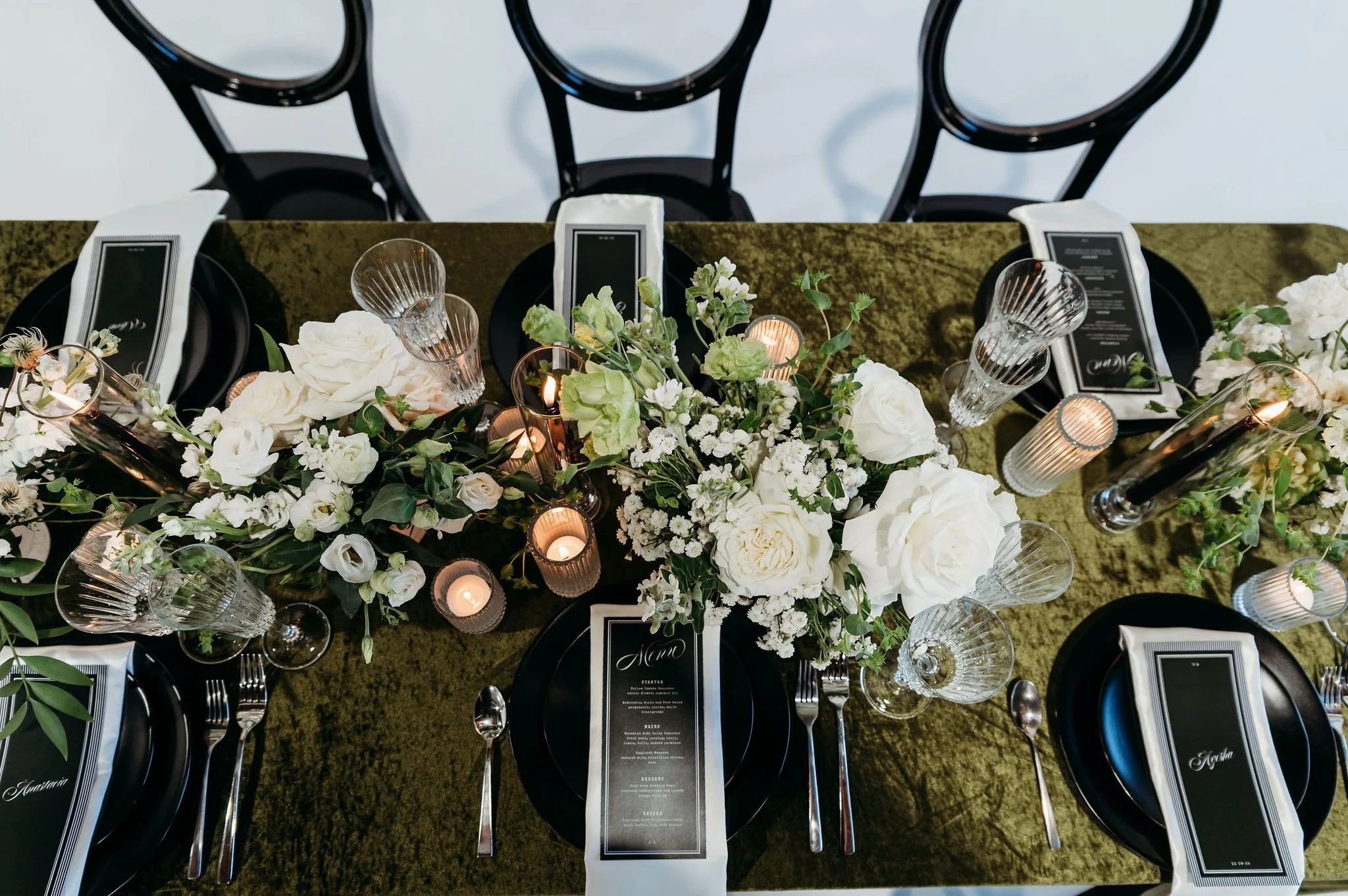 Elegant table setting with white floral centerpieces, candles, black plates, and water glasses, arranged for a formal event or dinner.