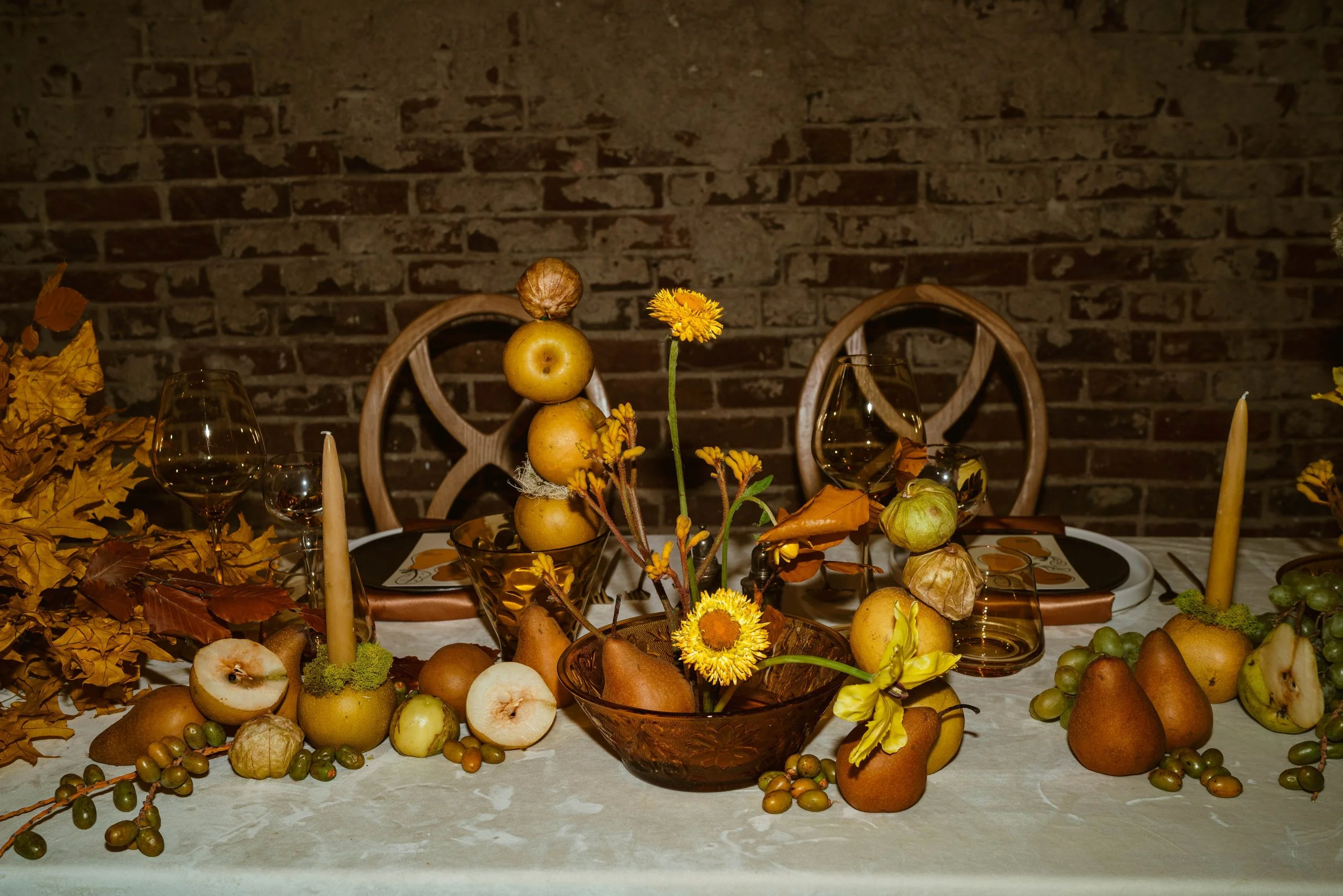 A dining table decorated with autumn-themed items including pears, apples, grapes, yellow flowers, dried leaves, candles, and glassware, set against a exposed brick wall.