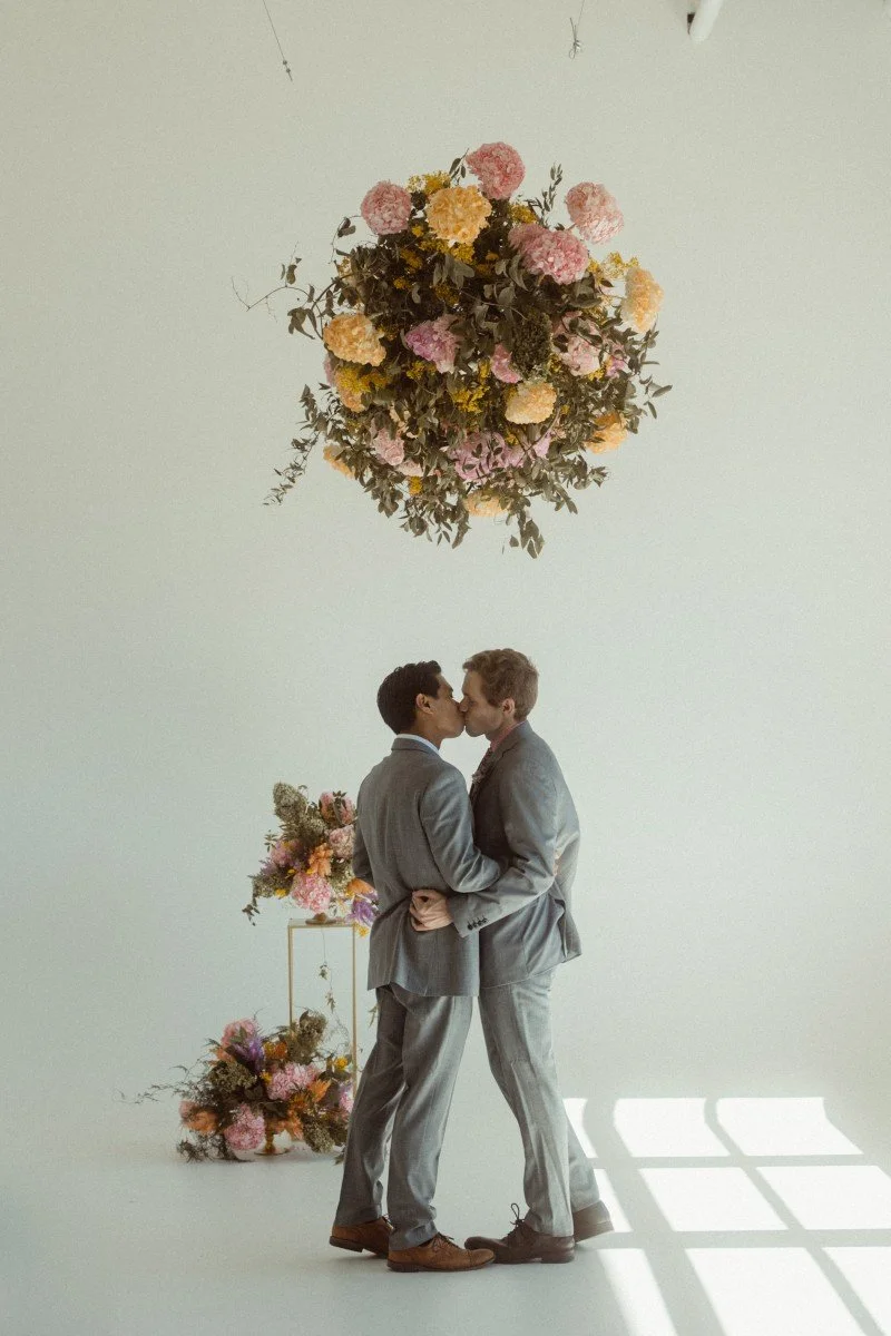 Two men wearing gray suits are kissing in a minimalist setting with a large hanging flower arrangement overhead and a smaller flower bouquet on a stand in the background.