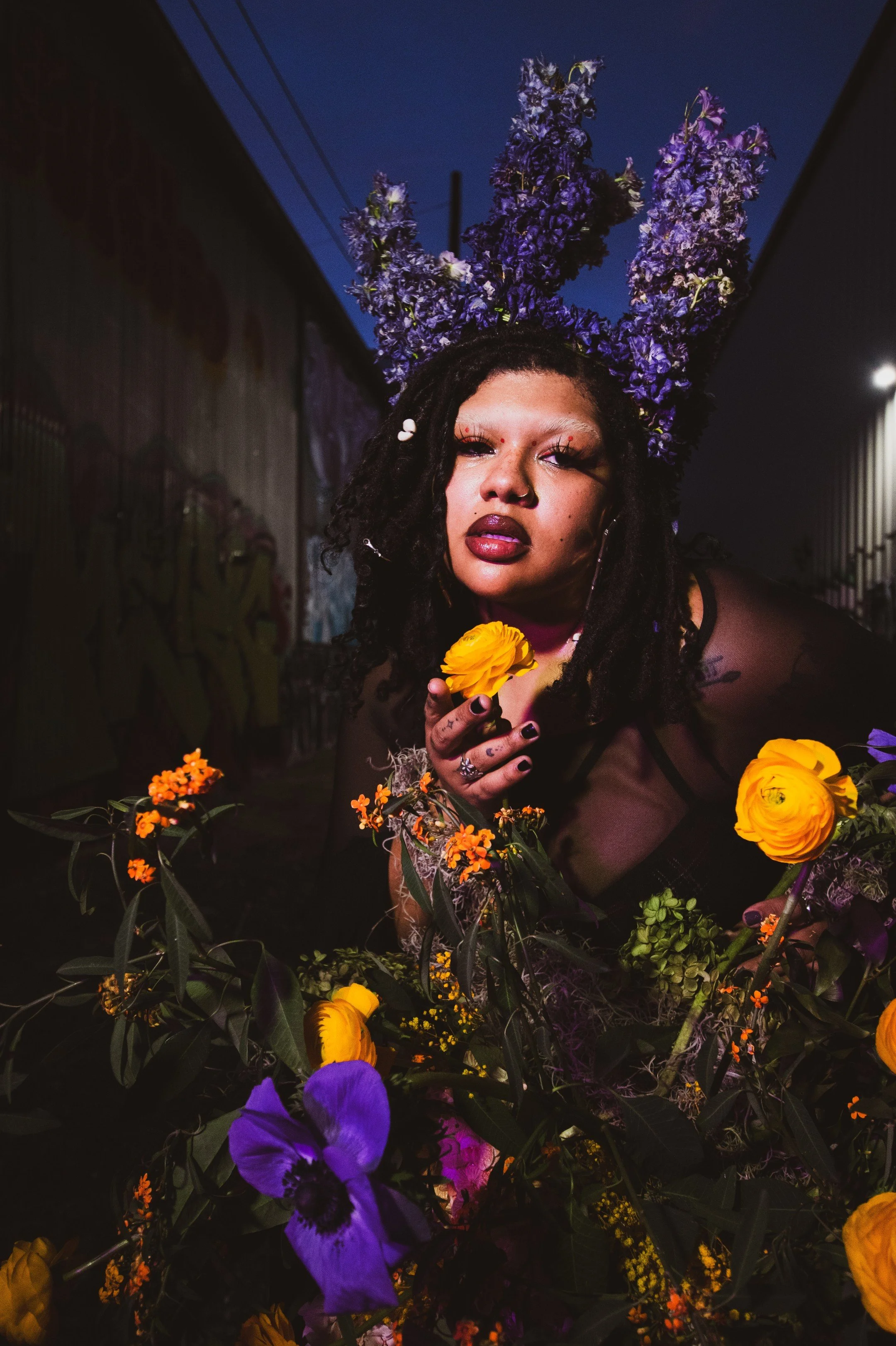 A woman with dark curly hair and bold makeup, including dark lipstick and decorated eyelashes, wears a headpiece made of large purple flowers. She is holding a yellow flower close to her face and is surrounded by an arrangement of yellow, purple, and orange flowers in a dark, urban alleyway at night.